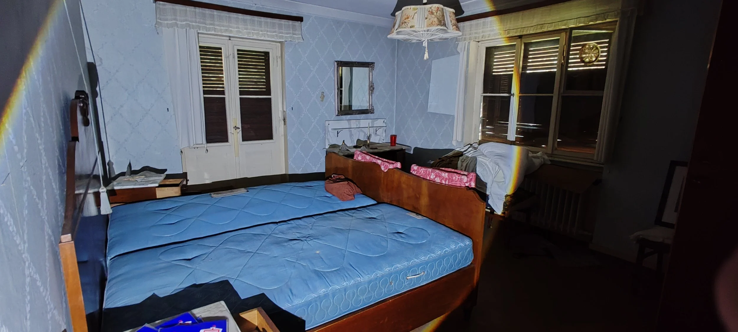 Ein unordentliches Schlafzimmer mit einem Doppelbett, einem Einzelbett, mehreren Fenstern und schäbigen Vorhängen, chaotischer Einrichtung und verschiedenen Gegenständen auf den Oberflächen.