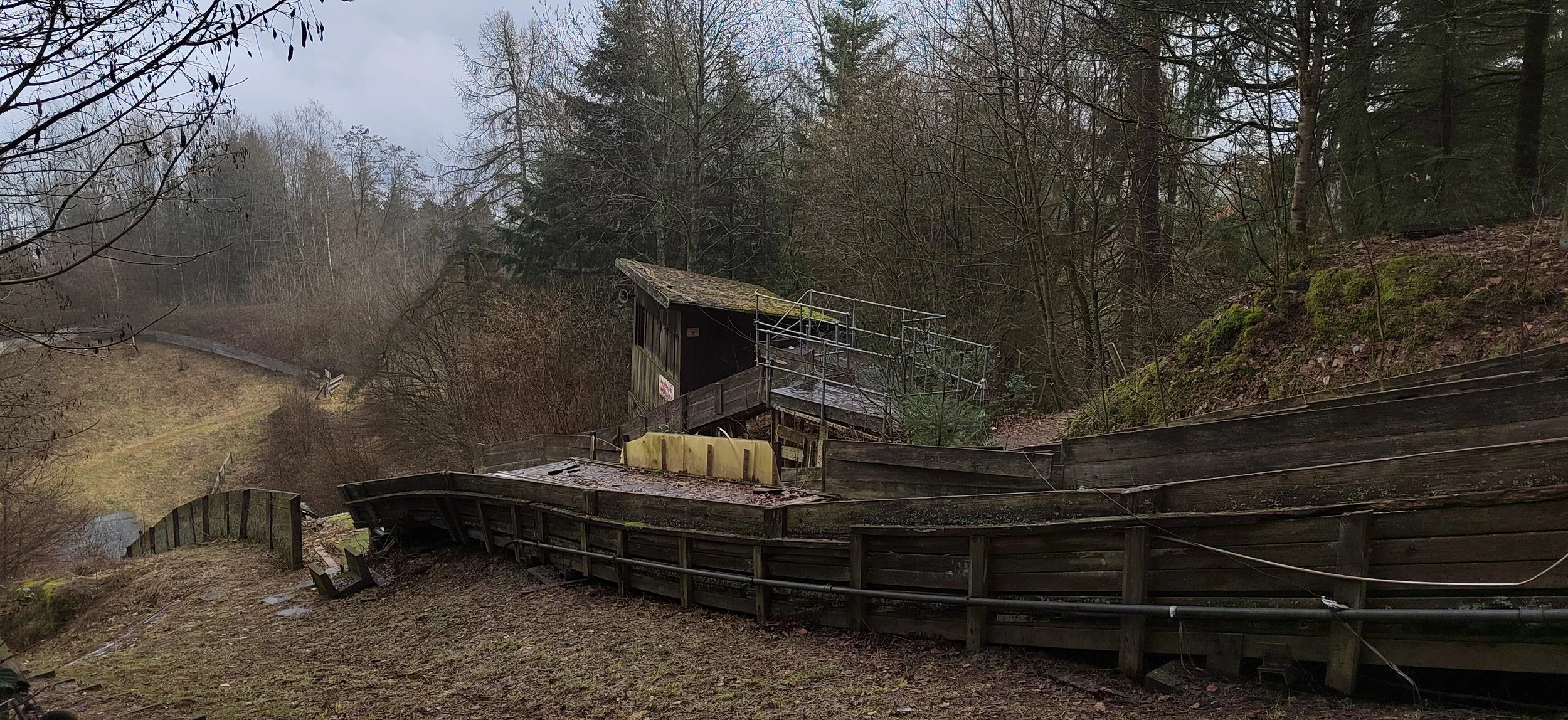 Ein verfallenes und zerfallenes Holzgebäude auf einem Hügel, umgeben von Bäumen und Sträuchern, bei bewölktem Himmel.