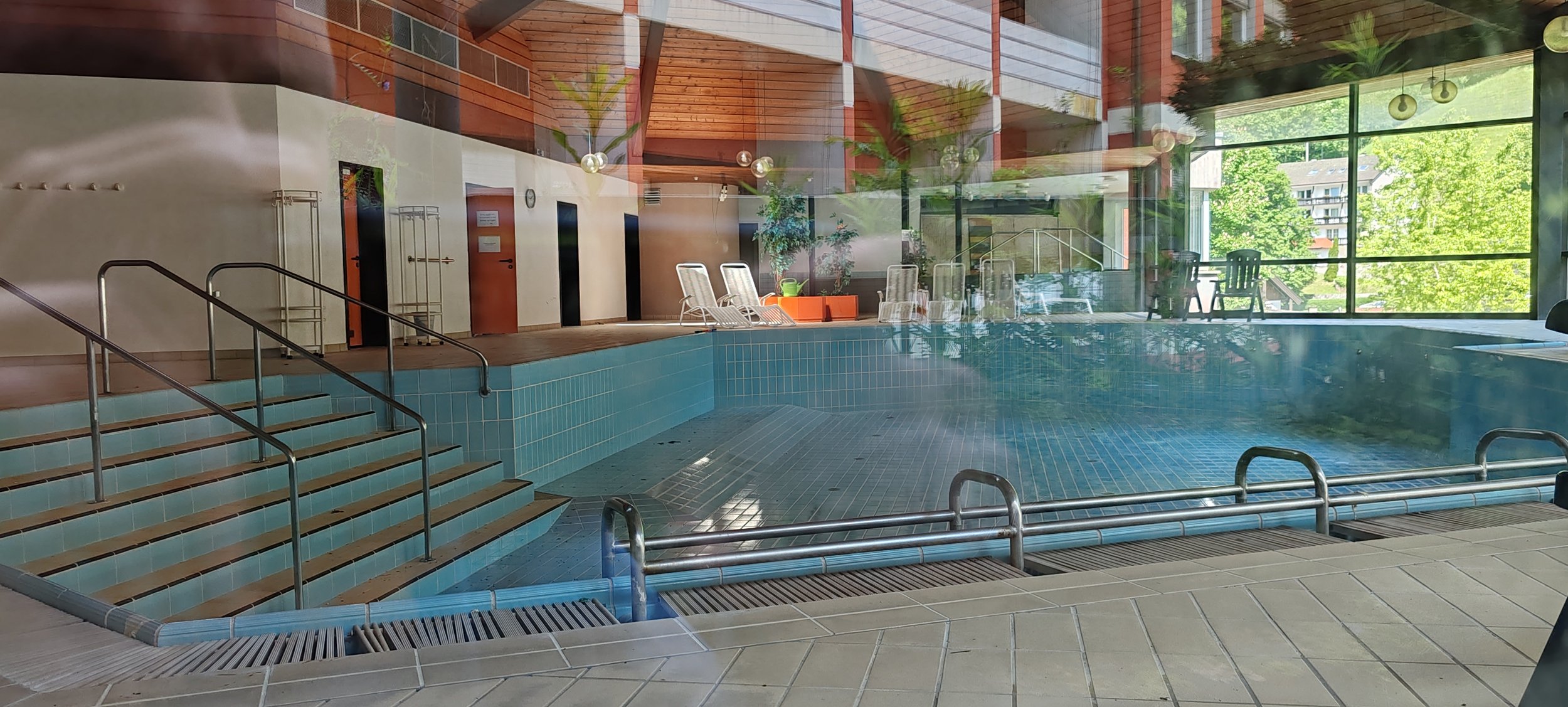 Innenbereich eines Schwimmbads mit Treppen, Liegen und großen Fensterfronten, durch die Bäume und Gebäude im Freien sichtbar sind.