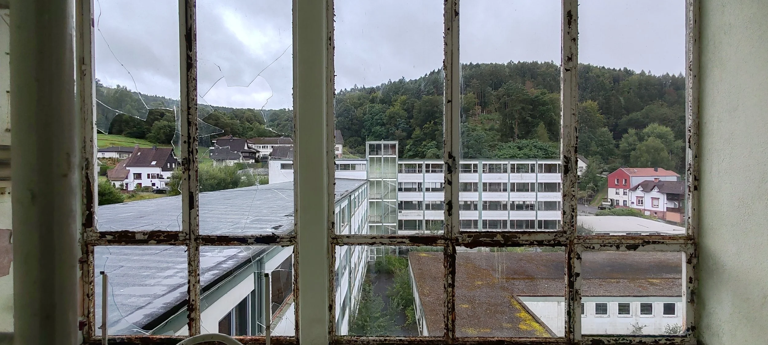 Blick durch ein altertümliches, zerbrochenes Fenster auf eine ländliche Wohngegend mit mehreren Häusern, einem mehrstöckigen modernen Gebäude und einer bewaldeten Hügelkette im Hintergrund, alles bei wolkigem Himmel.