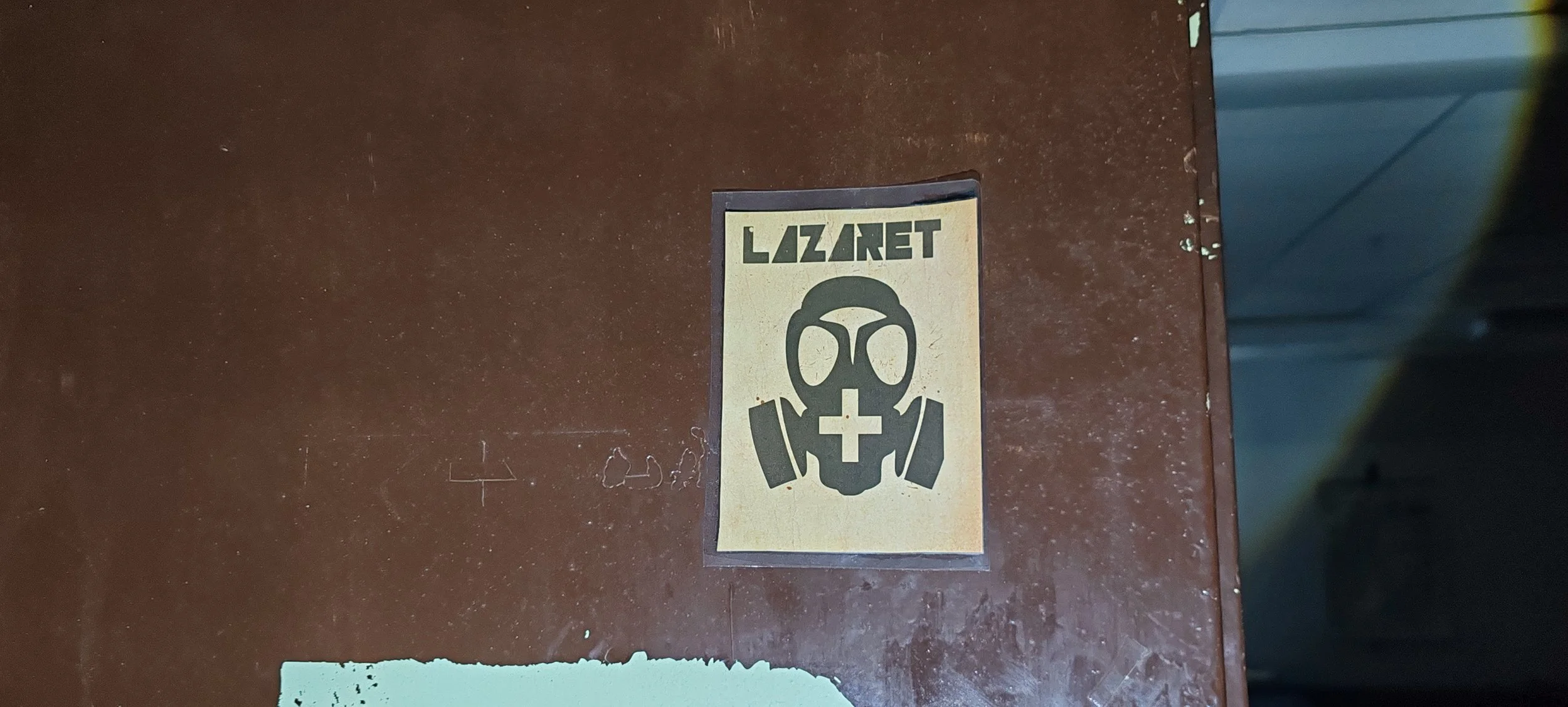 Aufkleber mit dem Text "LAZARET" und einem Symbol eines Gasmaske mit einem Kreuz in der Mitte, auf einer braunen Oberfläche.