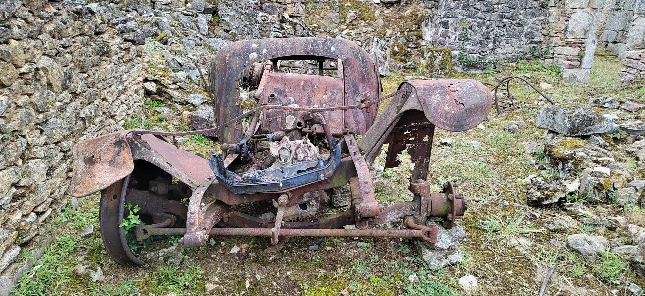 Verrosteter, verfallener Fahrzeugrahmen auf einer verschlungenen Grünfläche, umgeben von Steinen und Mauern.