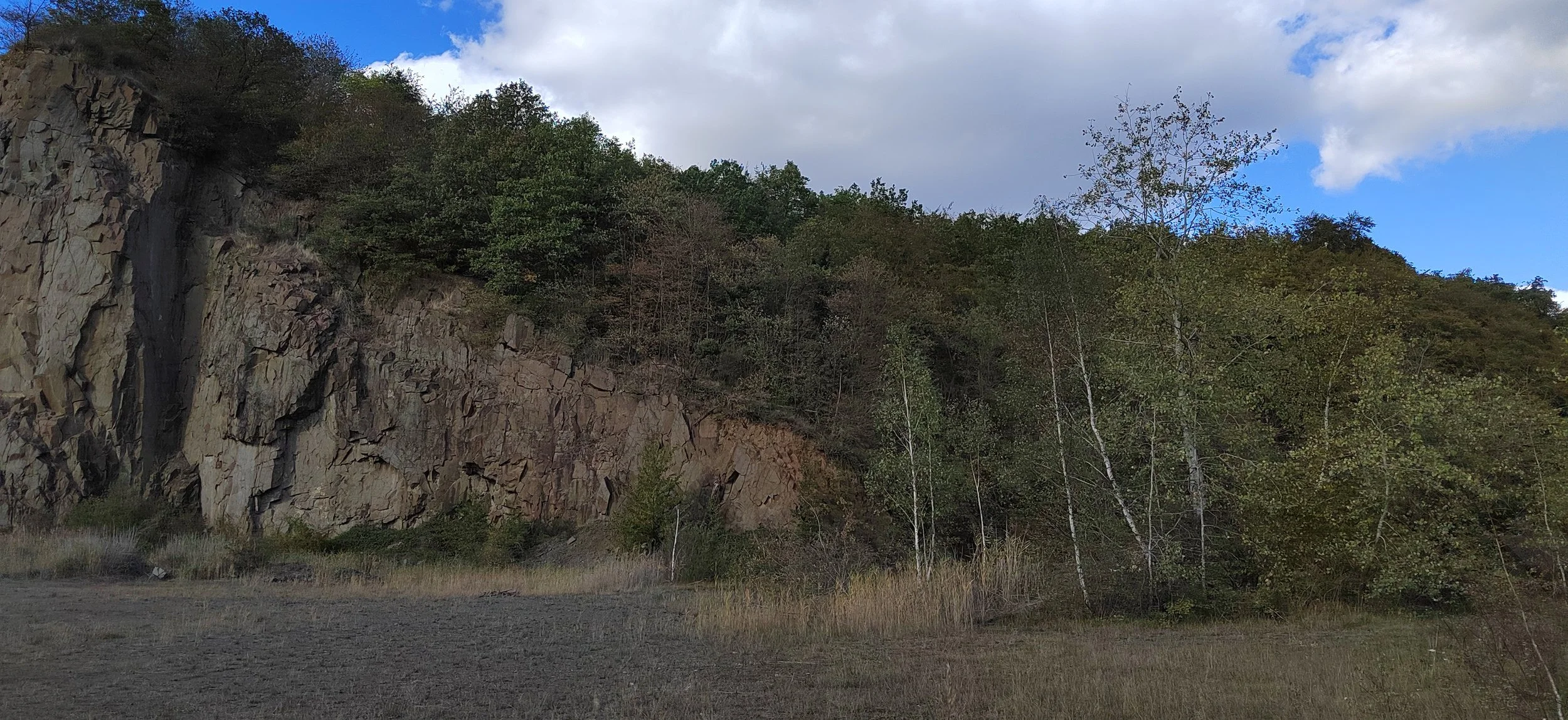 Hügel mit Felswand und Bäumen, im Vordergrund trockener Gras, Himmel mit Wolken und blauer Lücke.