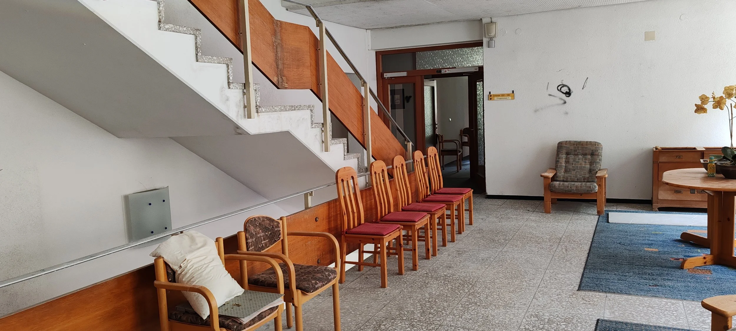 Ein Wartezimmer mit mehreren Holzstühlen, einer gepolsterten Sessel, einem Bücherregal mit Blume und einer Tür im Hintergrund.