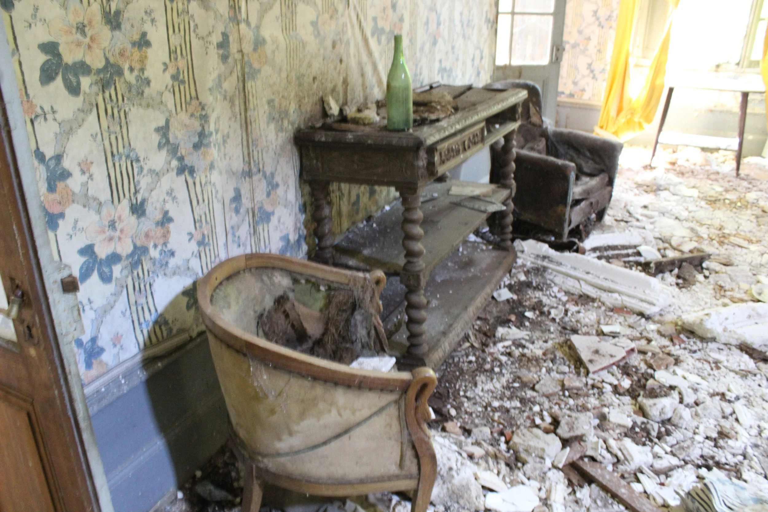 Verfallene, verstaubte Wohnzimmer mit zerbrochenem Mobiliar, Schutt und Staub. Ein alter Sessel, ein Regal mit einer grünen Flasche auf der Spitze, eine zerfallene Couch, alles ist von Staub und Trümmern bedeckt. Das Fenster im Hintergrund lässt lich