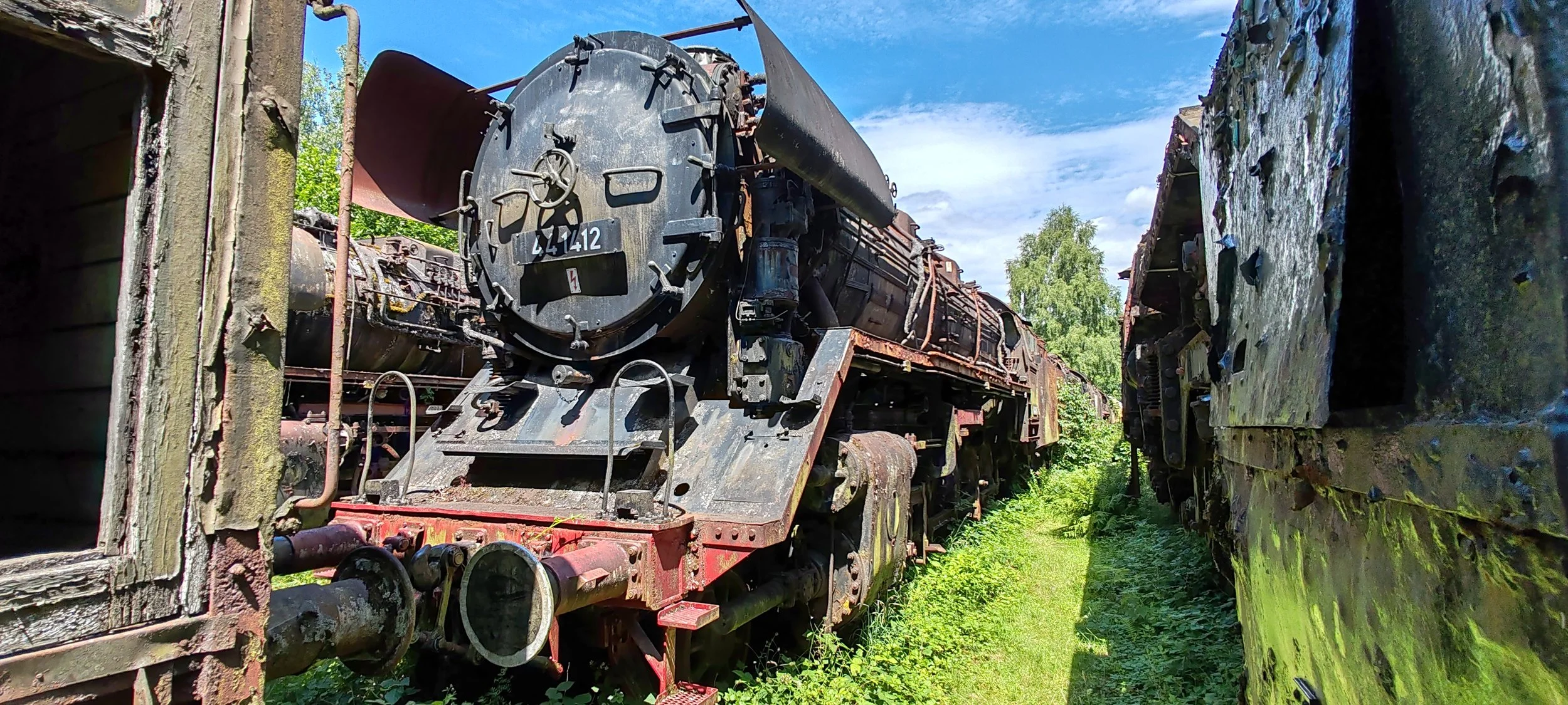 Verrostete, alte Dampflokomotive zwischen verfallenem Bahngleis und grüner Natur, unter blauem Himmel.