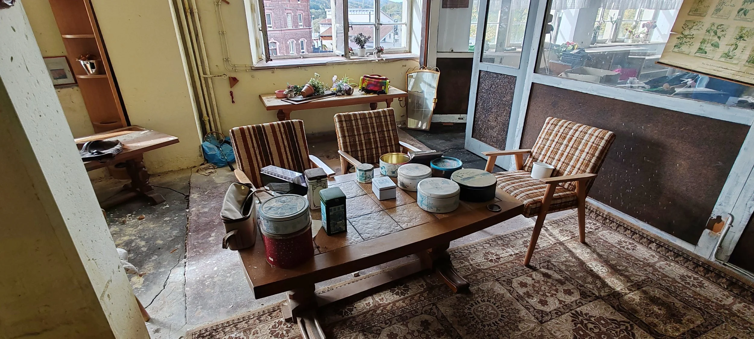 Innenraum eines abgelegenen Zimmers mit einem großen Holztisch, umgeben von acht Stühlen mit karierten Sitzbezügen. Auf dem Tisch befinden sich verschiedene alte Dosen und eine Tasche. Im Hintergrund sind offene Fenster, durch die man auf benachbarte