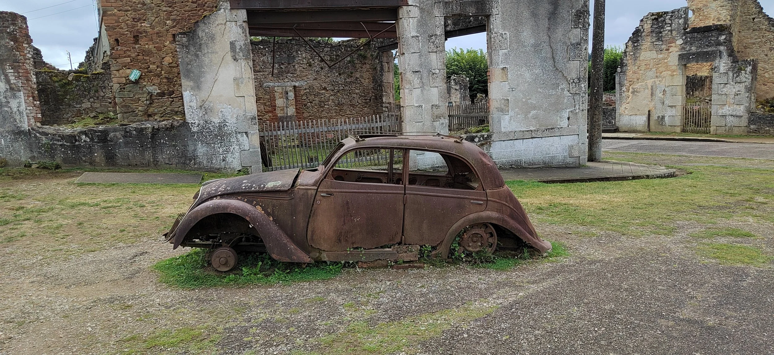 Verrosteter, alter Auto-Kadaver steht auf einem Feld vor den Ruinen eines alten Gebäudes.