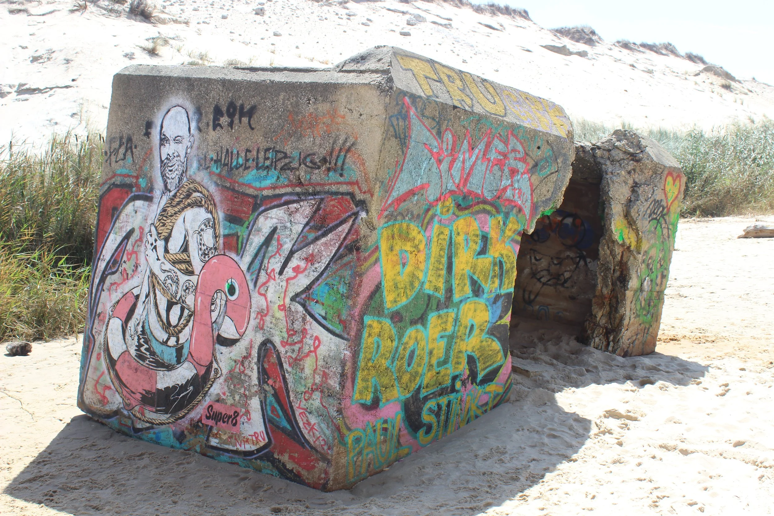 Graffiti-Kunstwerk auf einem Betonblock am Strand, mit Skizzen, bunten Schriftzügen und einem Menschenporträt, umgeben von Sand und Strandvegetation.