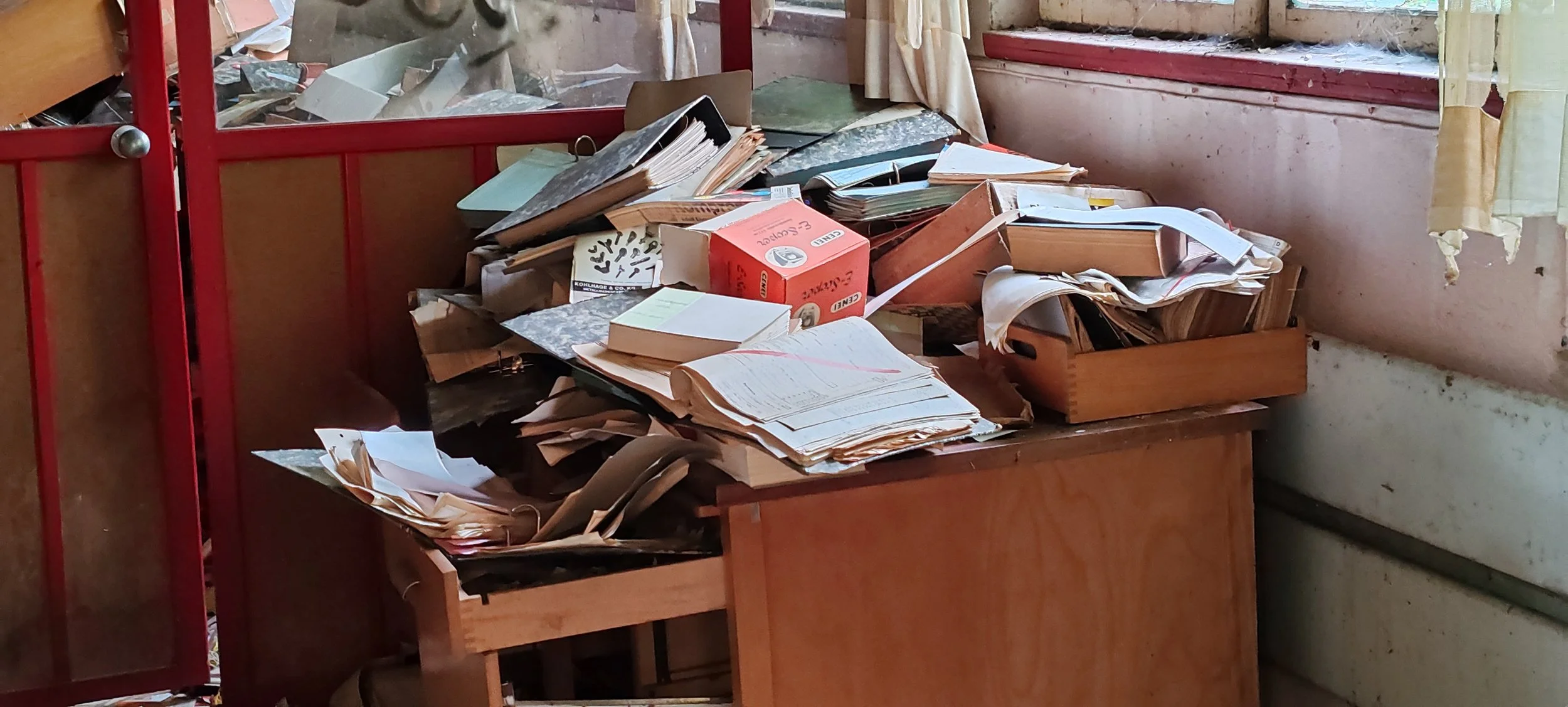 Ein chaotischer Schreibtisch mit Papierstapeln, Ordnern und einem Boxenstapel in einem vernachlässigten Raum, neben einem Fenster mit zerfetzter Vorhang.