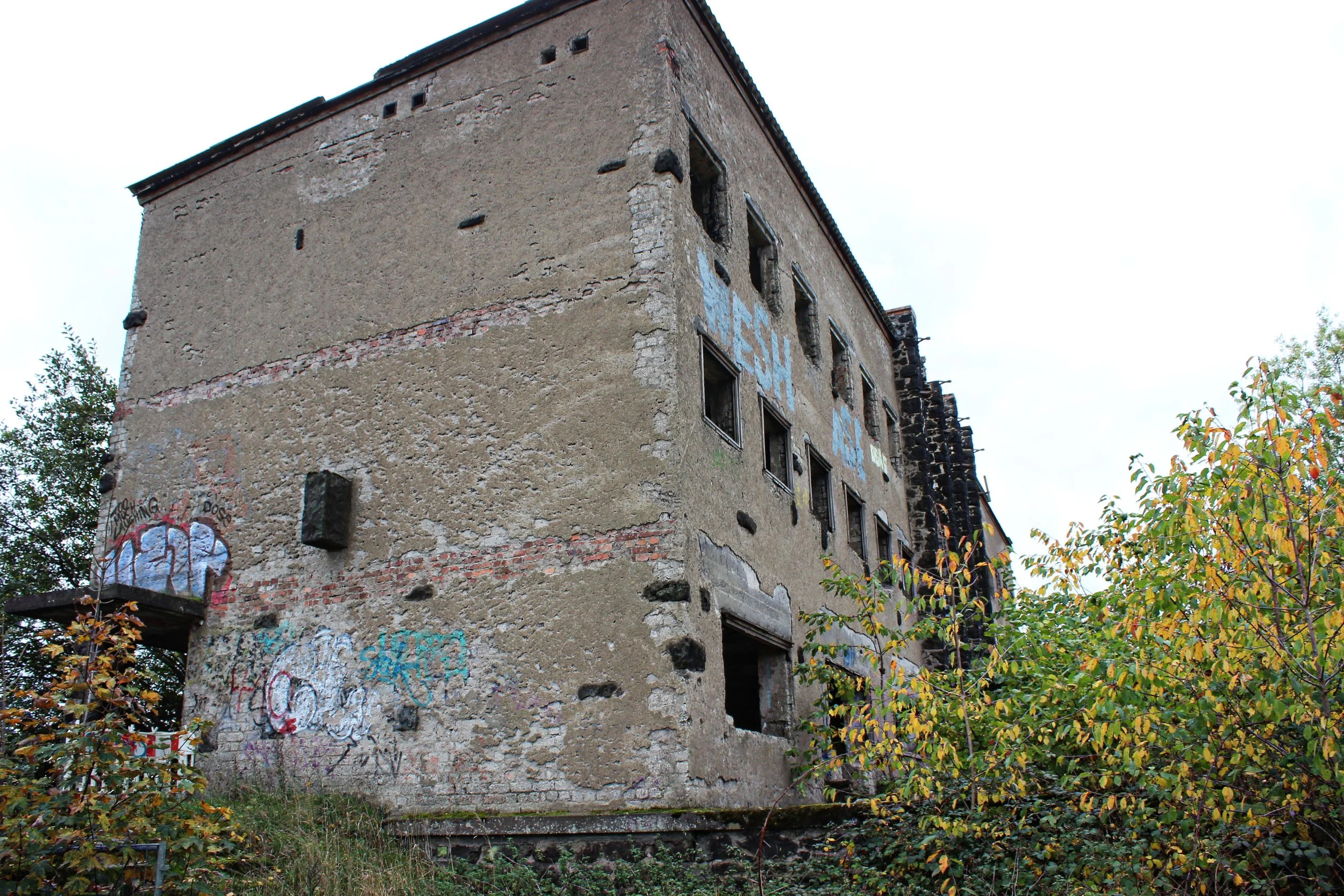 Ein verfallenes, bröckelndes Gebäude mit mehreren zerbrochenen Fenstern, teilweise mit Graffiti bemalt, umgeben von Vegetation.