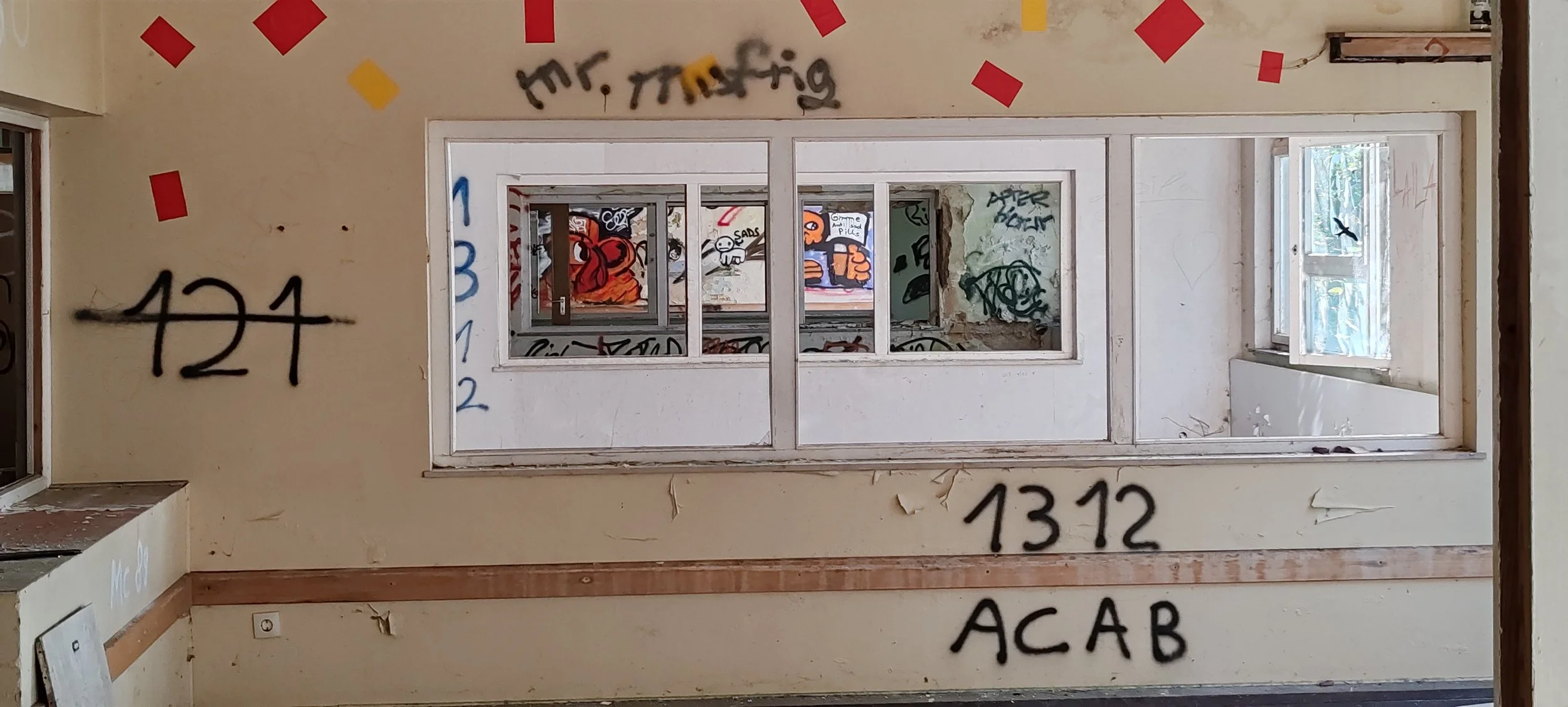 Ein verlassenes, Graffiti-verschmiertes Zimmer mit einer großen, zerbrochenen Fensteröffnung, durch die ein weiteres graffitibestempeltes Raum sichtbar ist.