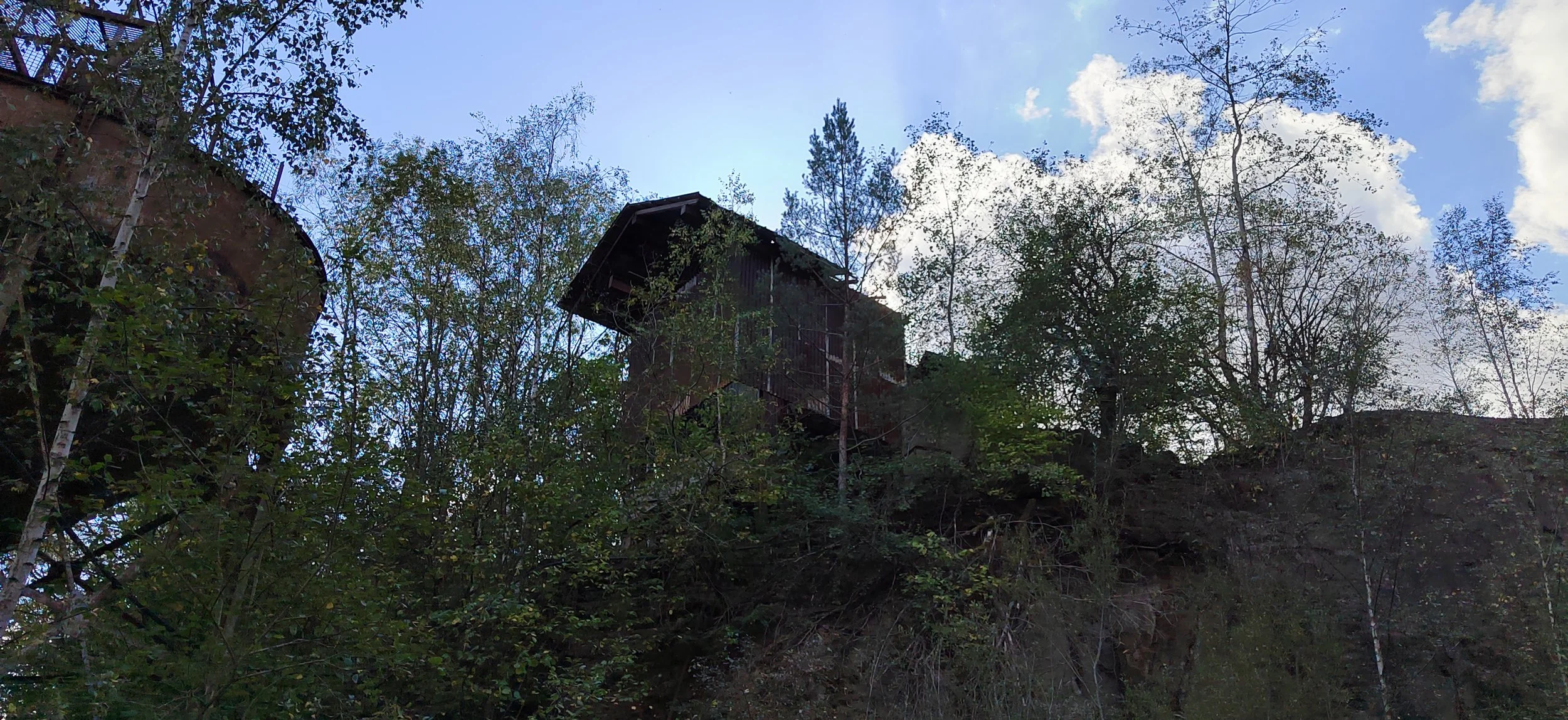 Ein Baumhaus auf einem Hügel, umgeben von Bäumen mit blauem Himmel und Wolken im Hintergrund.