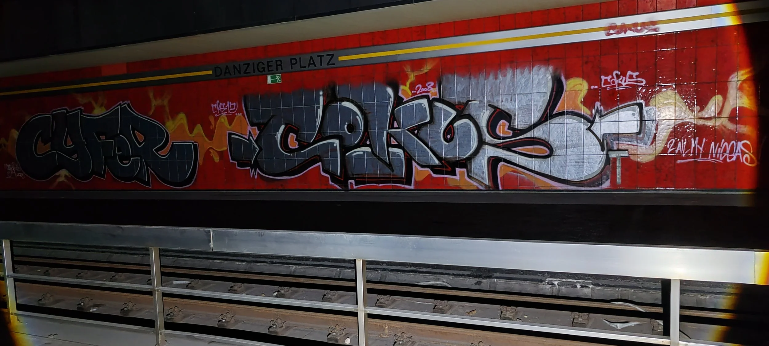Graffiti an der Wand im U-Bahnhof Danziger Platz, in Schwarz, Hellblau, Hellorange, und Weiß auf rotem Hintergrund.