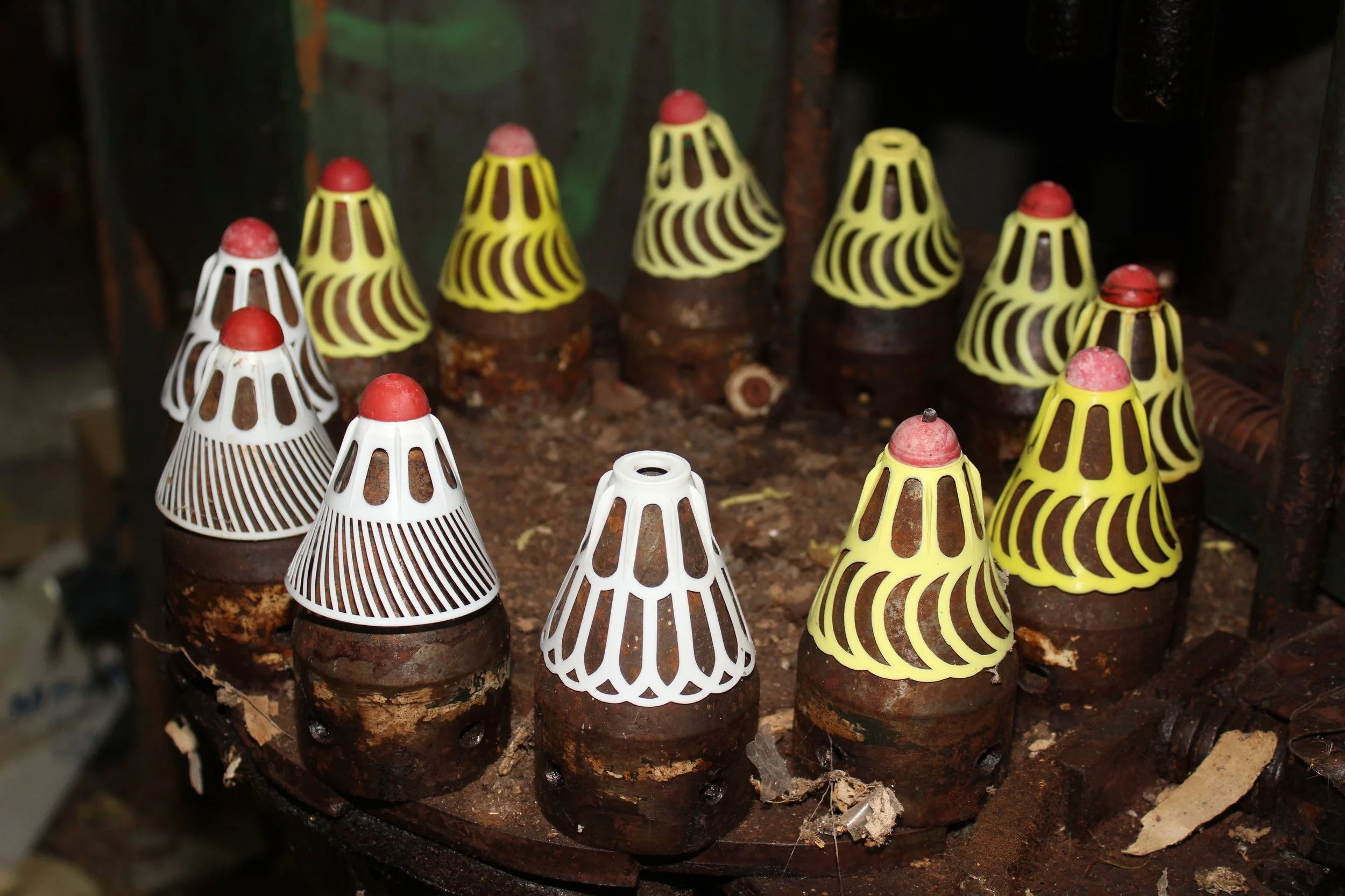 Mehrere traditionelle Fliegenklatschen aus Holz und Kunststoff, in Weiß und Gelb, mit rotgrünen Griffen, liegen auf einem rustikalen Holzblock, umgeben von Erde und Spuren von Holzspänen.