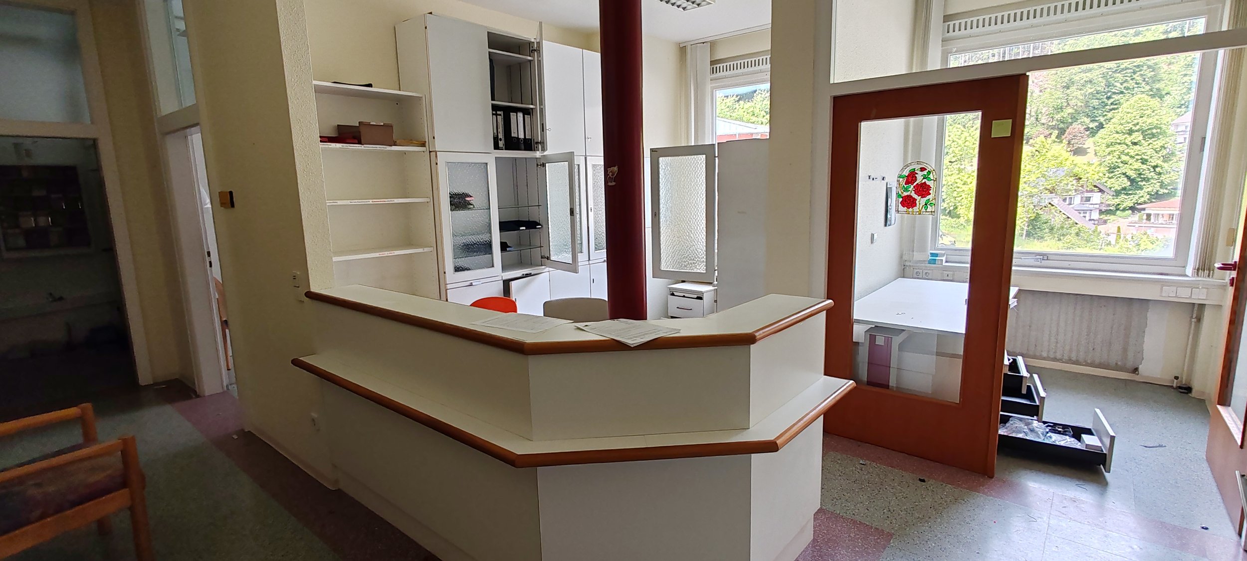 Leere Rezeption in einem geschlossenen Büro mit großen Fenstern, einem Tresen und einem Stuhl. Draußen sind Bäume und Gebäude sichtbar.