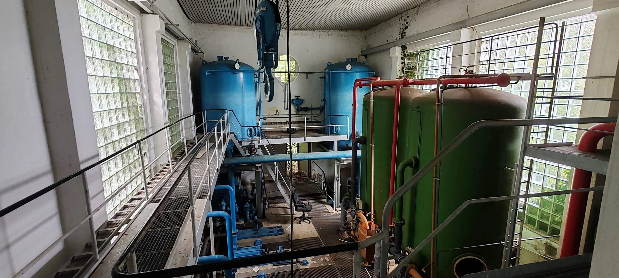 Ein industrieller Raum mit großen blauen und grünen Tanks, Rohrleitungen und einer Hebebühne.