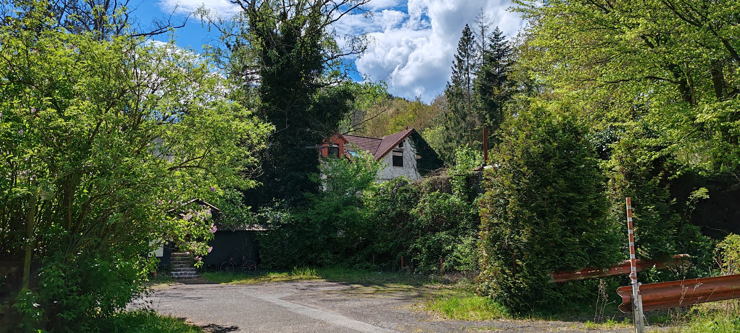 Ein Haus mit rotem Ziegeldach, umgeben von Bäumen und Sträuchern, auf einer kurvigen Straße im Grünen, bei sonnigem Wetter mit einigen Wolken am Himmel.