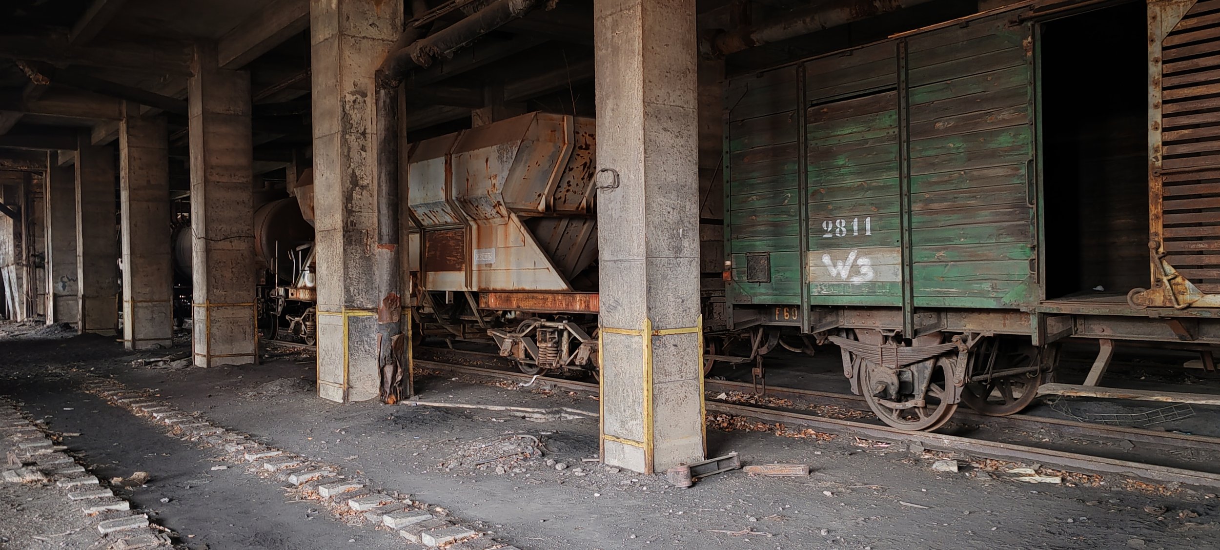 Verfallene und verrostete alte Güterwagons in einer verlassenen Bahnstation, umgeben von Staub und Schutt.
