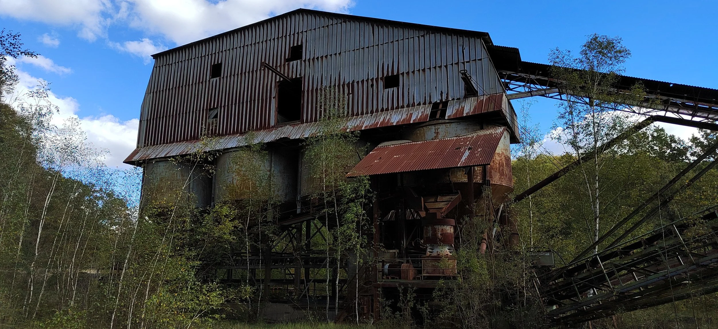 Verrostete, verlassene Fabrik mit Metallgebäude und Förderband, umgeben von Bäumen, under wolkigem Himmel.