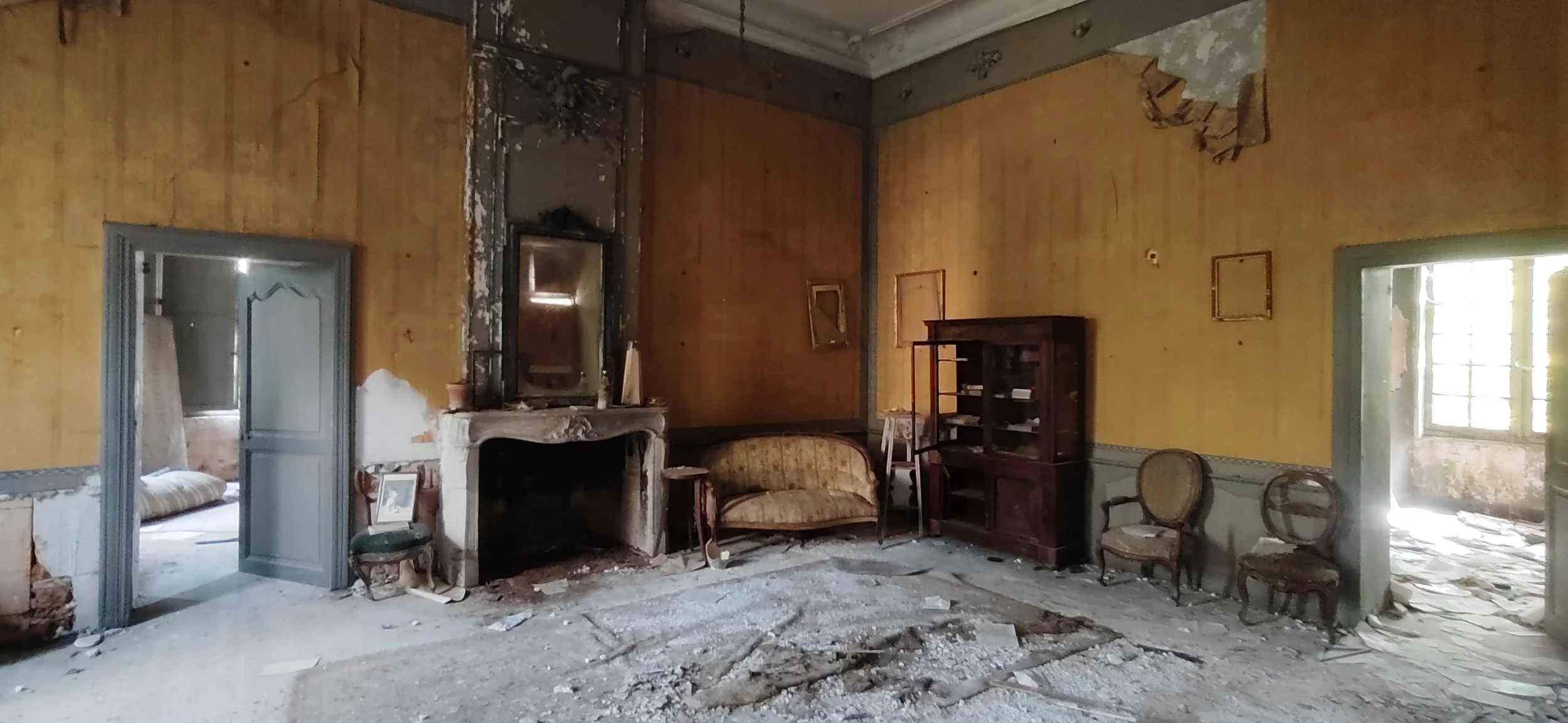 Ein verfallenes, verlassenes Wohnzimmer mit gelben Wänden, kaputtem Boden und antiken Möbeln wie Sofa, Stühlen, einem Kamin und einem Regal; das Zimmer ist schmutzig und zerfallen.