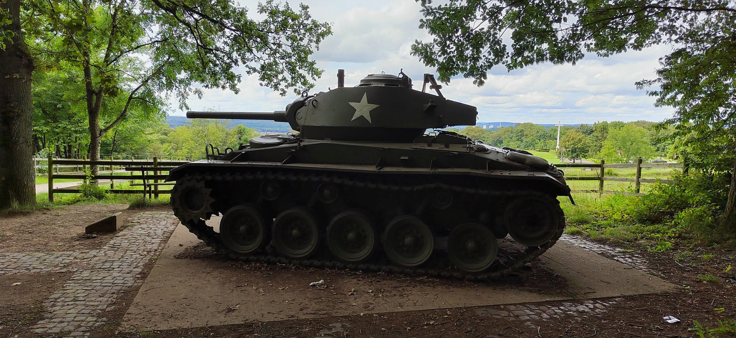 Ein alter Tank steht in einem Park, umgeben von Bäumen und einer Holzzäune, mit einer grünen Landschaft und einem Himmel mit Wolken im Hintergrund.