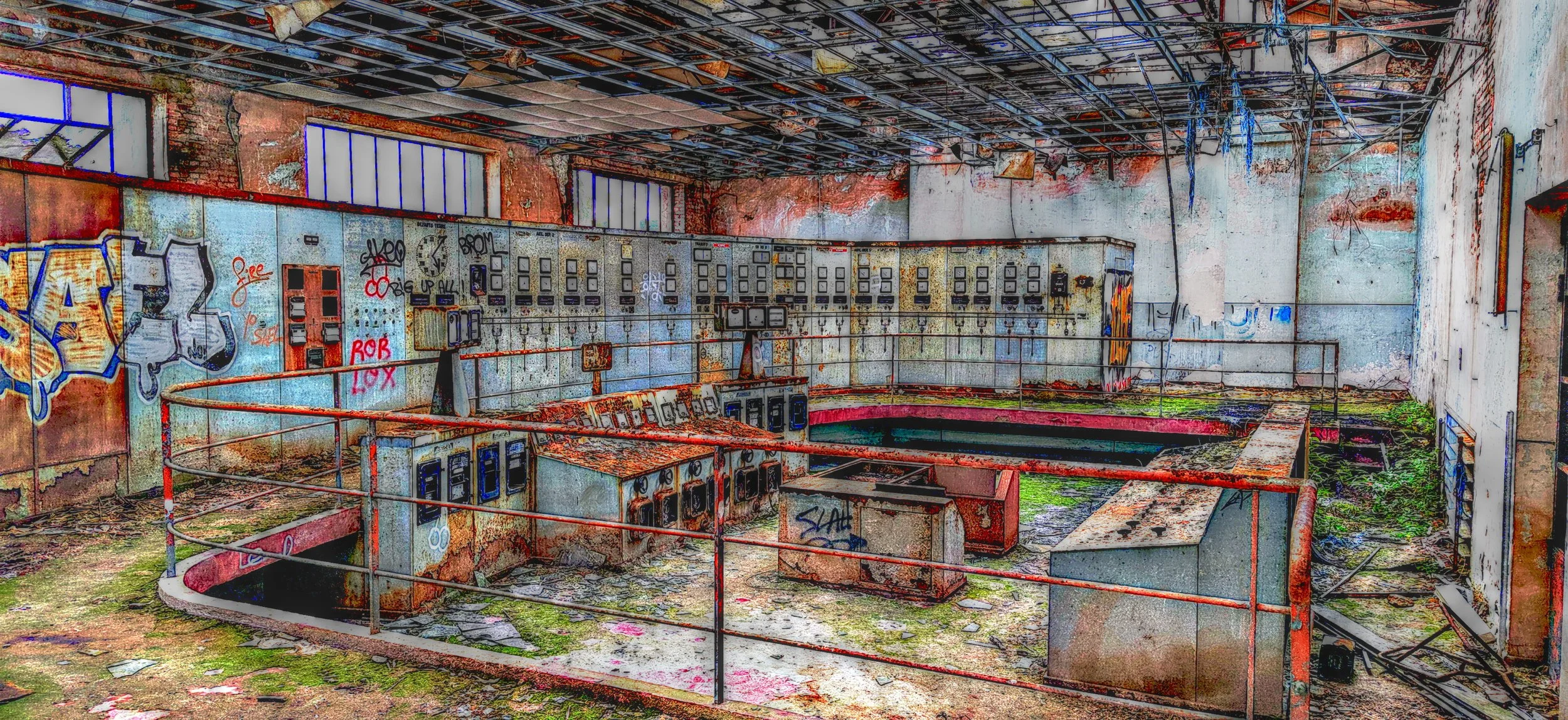 Verlassene, verfallene Industrieanlage mit Kontrollpulten, Graffiti an den Wänden, verwesendem Boden mit Moos und Schmutz, aufgerissene Decke mit sichtbaren Trägern.
