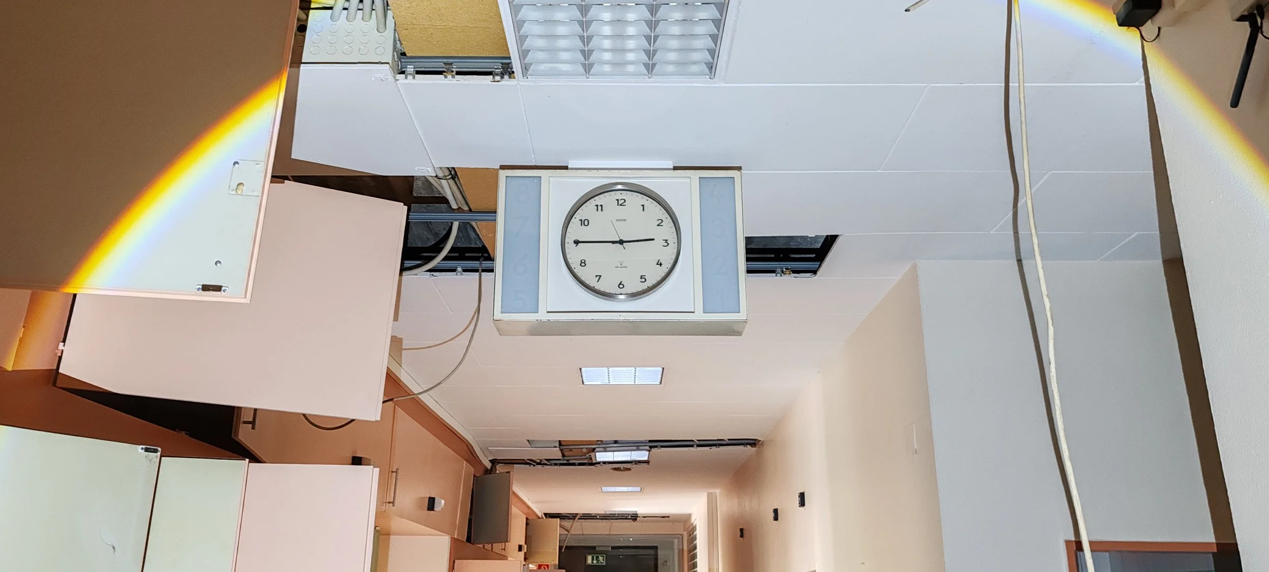 Ein Wanduhren in einem Flur im Inneren eines Gebäudes, mit weißen Wänden und Decken. Der Flur ist lang und hell, mit einigen Türen an den Seiten.