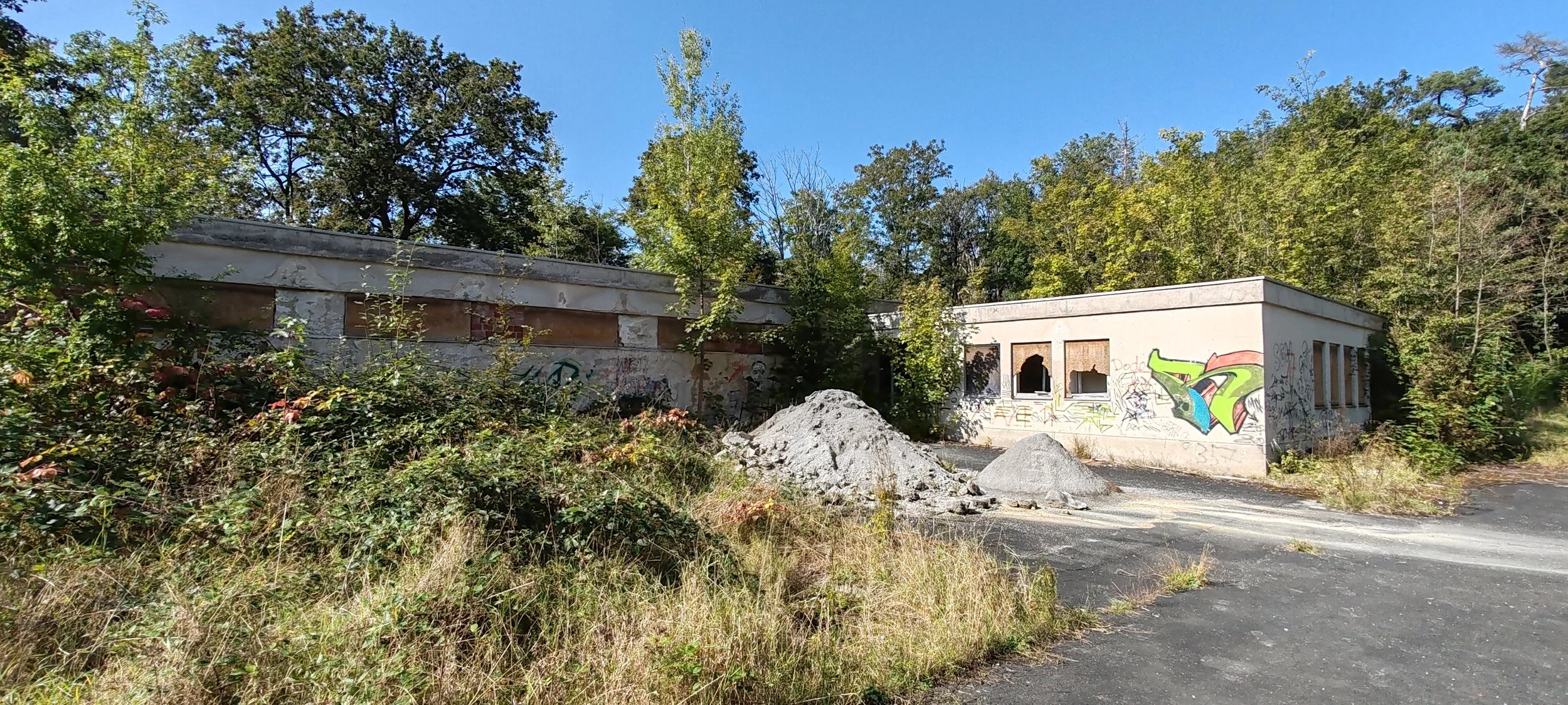 Verfallene Gebäude mit Graffiti, überwuchert von Pflanzen, mit Massen an Sand und Steinen im Vordergrund, umgeben von Bäumen bei sonnigem Himmel.