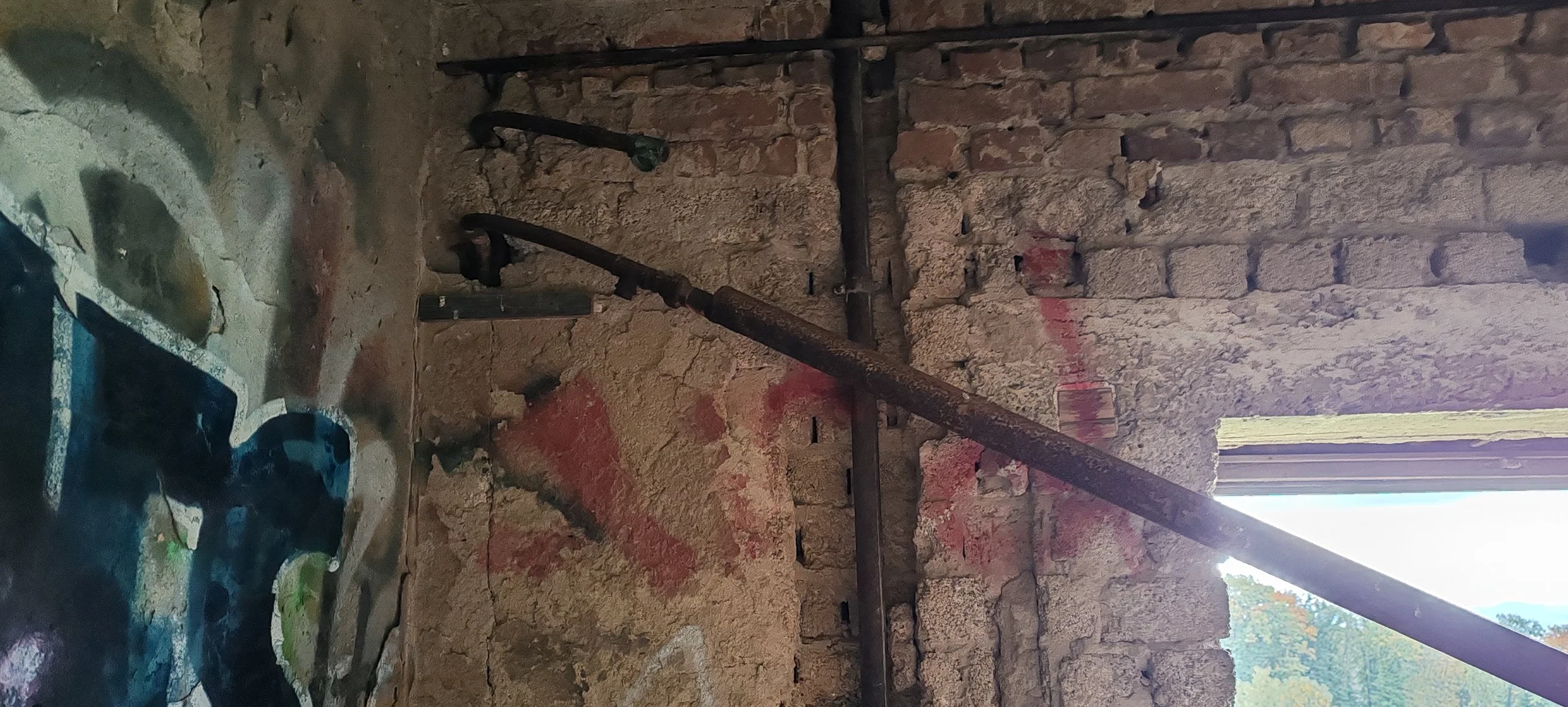 Alte, verrostete Wasserleitung an einer Steinausfachung in einer brüchigen Mauer, darunter eine bunte, ausgewaschene Wand mit Graffiti.