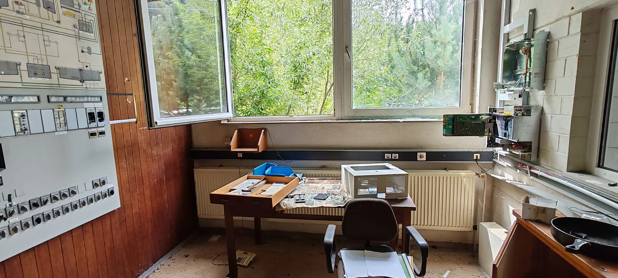 Innenraum eines veralteten Klassenzimmers mit großen Fenstern, alten Lehrmitteln, einem Tisch mit technischen Geräten, einem Bürostuhl und einem Schreibtisch, an dem verschiedene Gegenstände liegen.