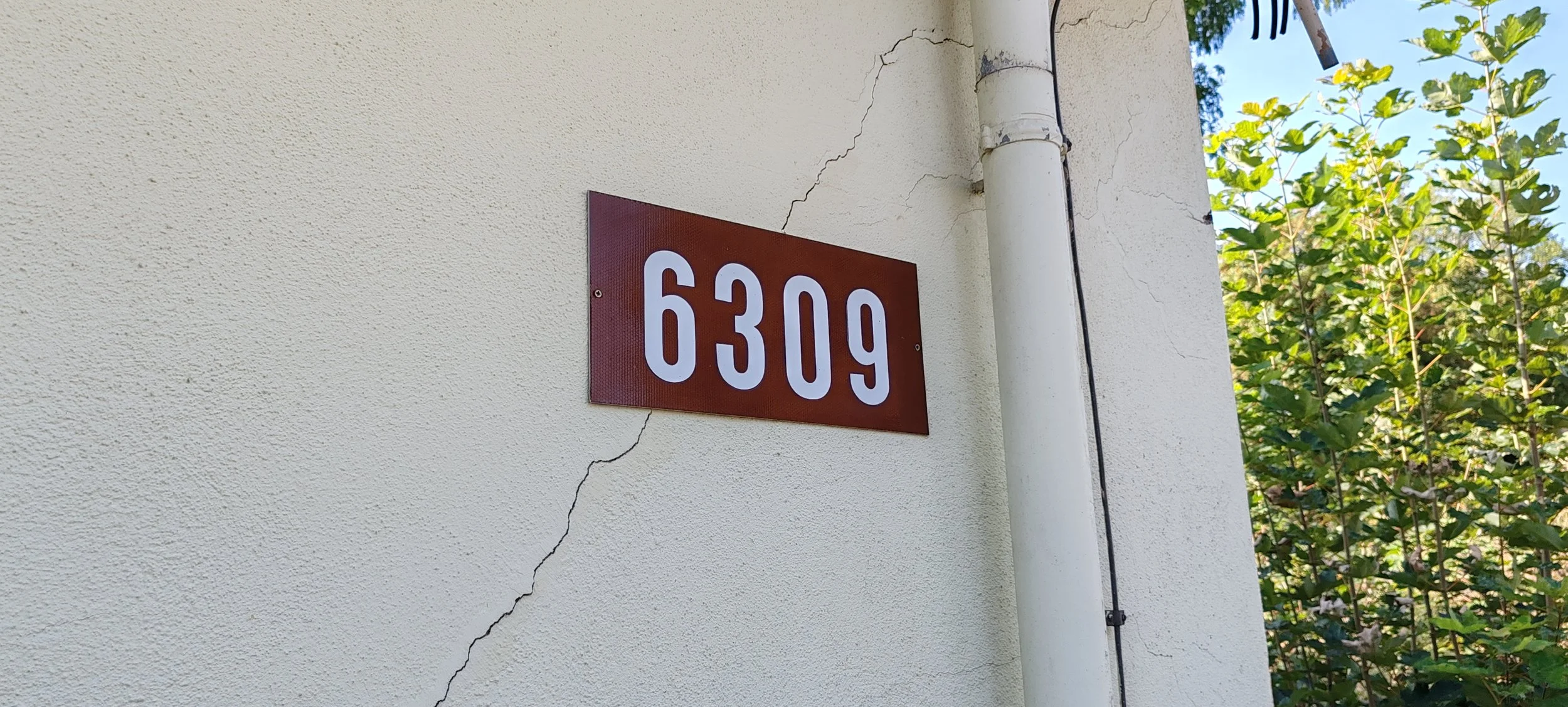 Hausnummer 6309 an einer cremefarbenen Wand mit Riss, neben einer weißen Regenrinne und grünem Laub im Hintergrund.