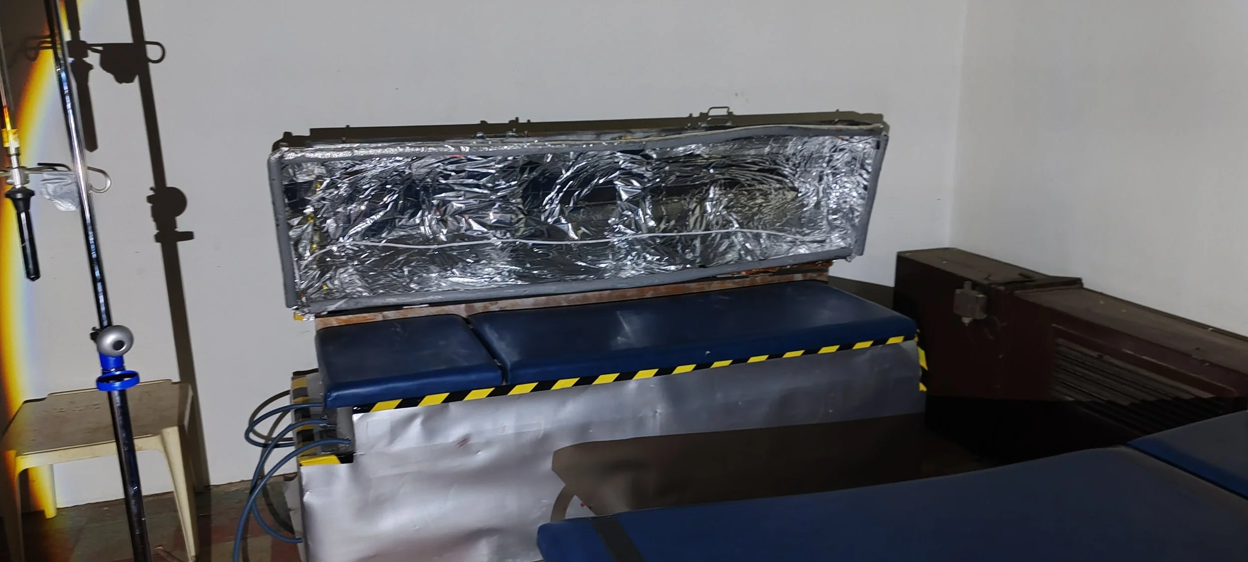 Ein medizinischer Untersuchungsraum mit einer Liege und mehreren medizinischen Geräten, darunter eine mit Aluminiumfolie bedeckte Maschine.