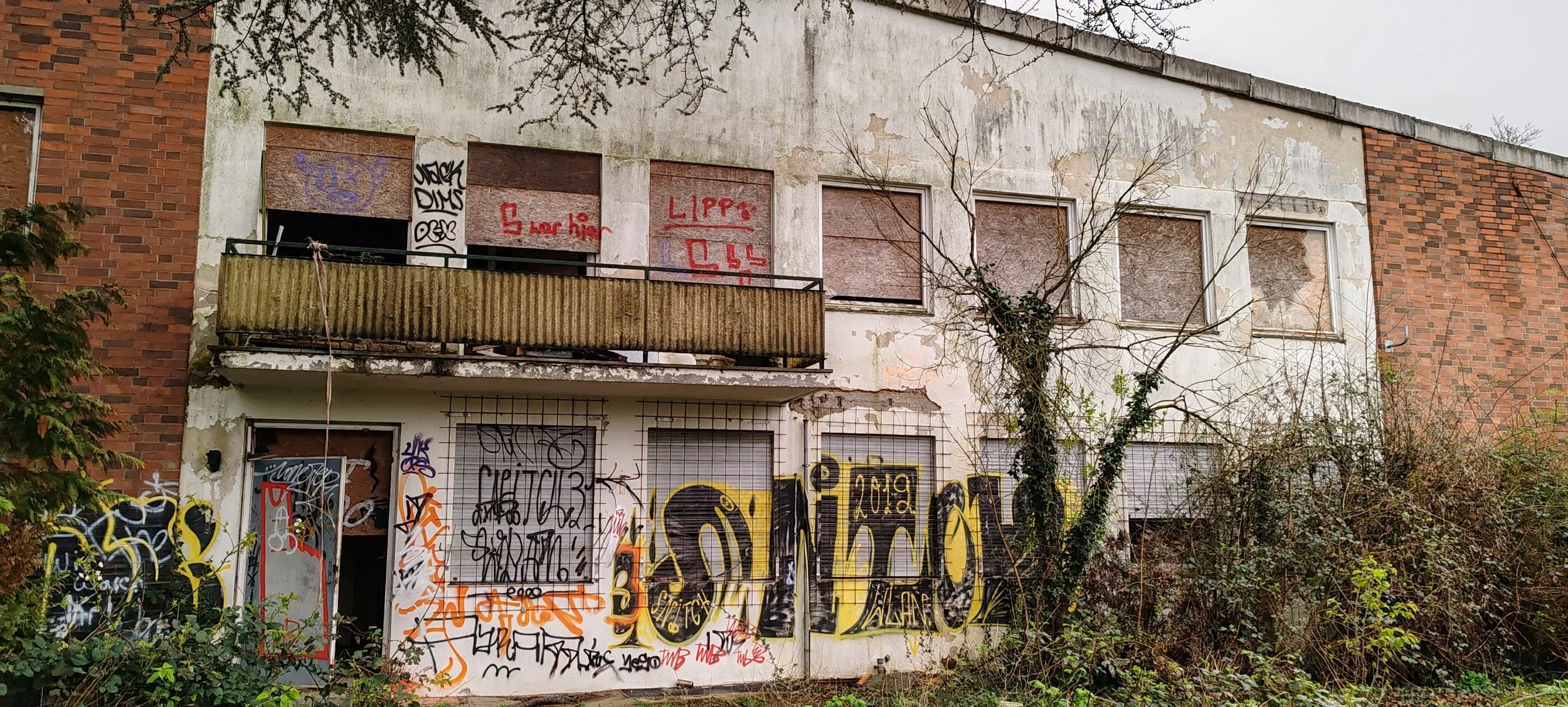 Ein verfallenes, von Graffiti bedecktes zweistöckiges Gebäude mit drei oberen Fenstern und einem Balkon. Das Gebäude ist teilweise verfallen, mit zerbröckelnder Fassade und abgestellten, zerbrochenen Fenstern. Um das Gebäude herum wachsen Sträucher u