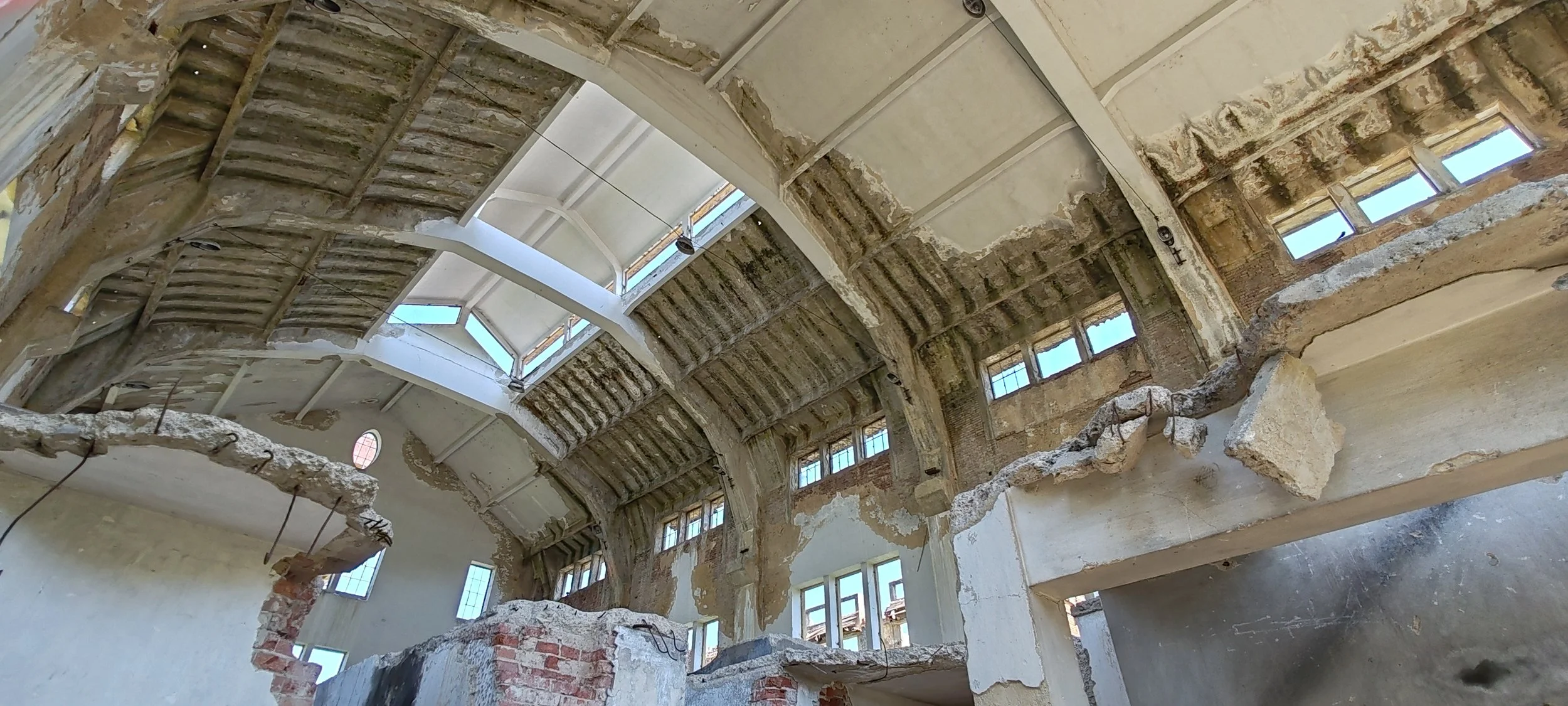Innenansicht eines alten Gebäudes in Sanierungsarbeiten, mit sichtbaren zerborstenen Wänden, großen Fenstern und teilweise offenem Dach, das Tageslicht hereinlässt.