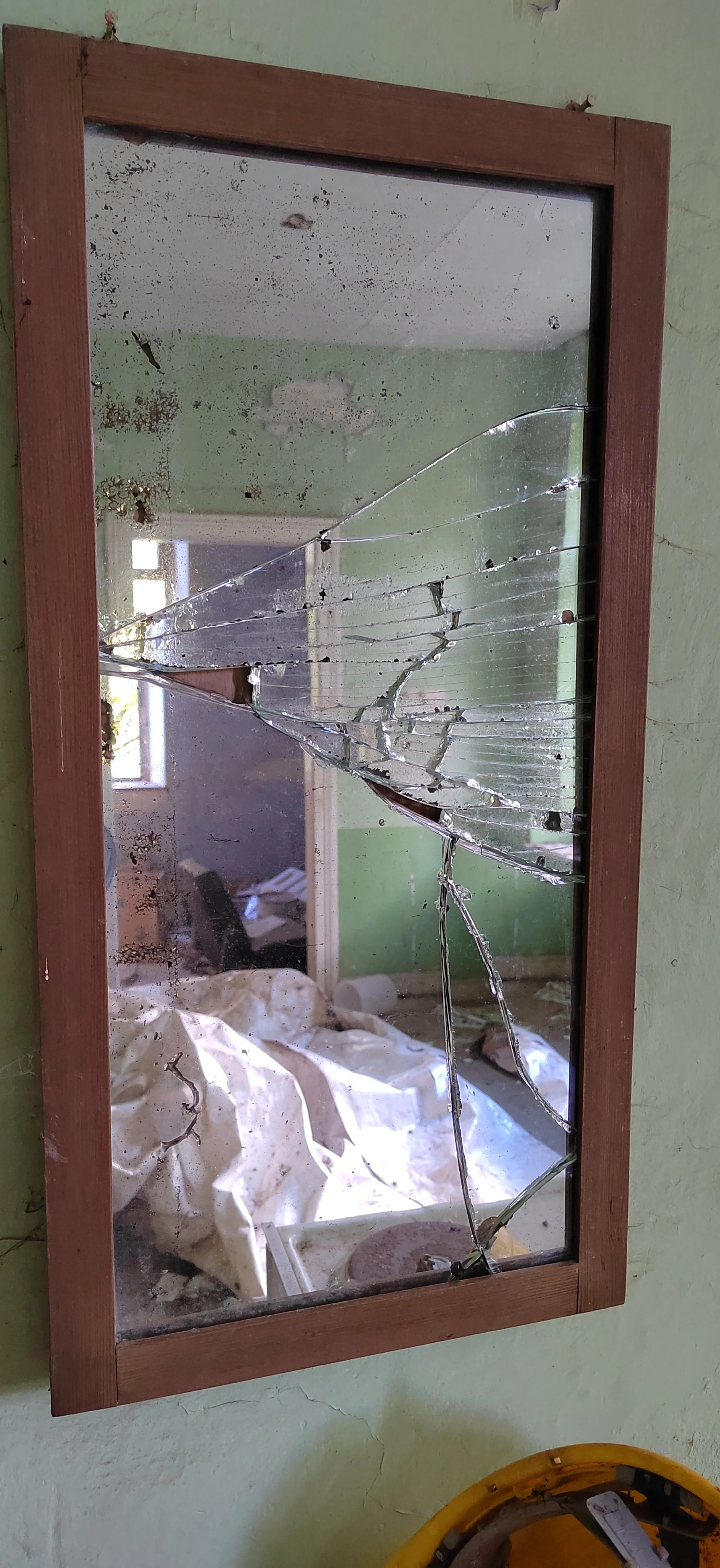 Zerbrochener Spiegel mit Rissen in einem Holzrahmen, gegenüber ein Raum mit grün gestrichener Wand, unordentlichem Bett und mehreren Gegenständen.