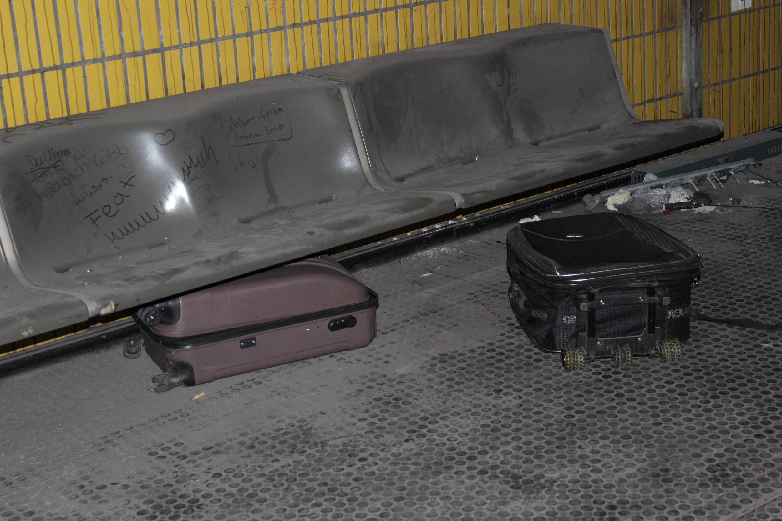 Leere wartende Parkbank mit Graffiti, daneben zwei kleine Gepäckstücke auf dem Boden, im Hintergrund eine gelbe Wand.