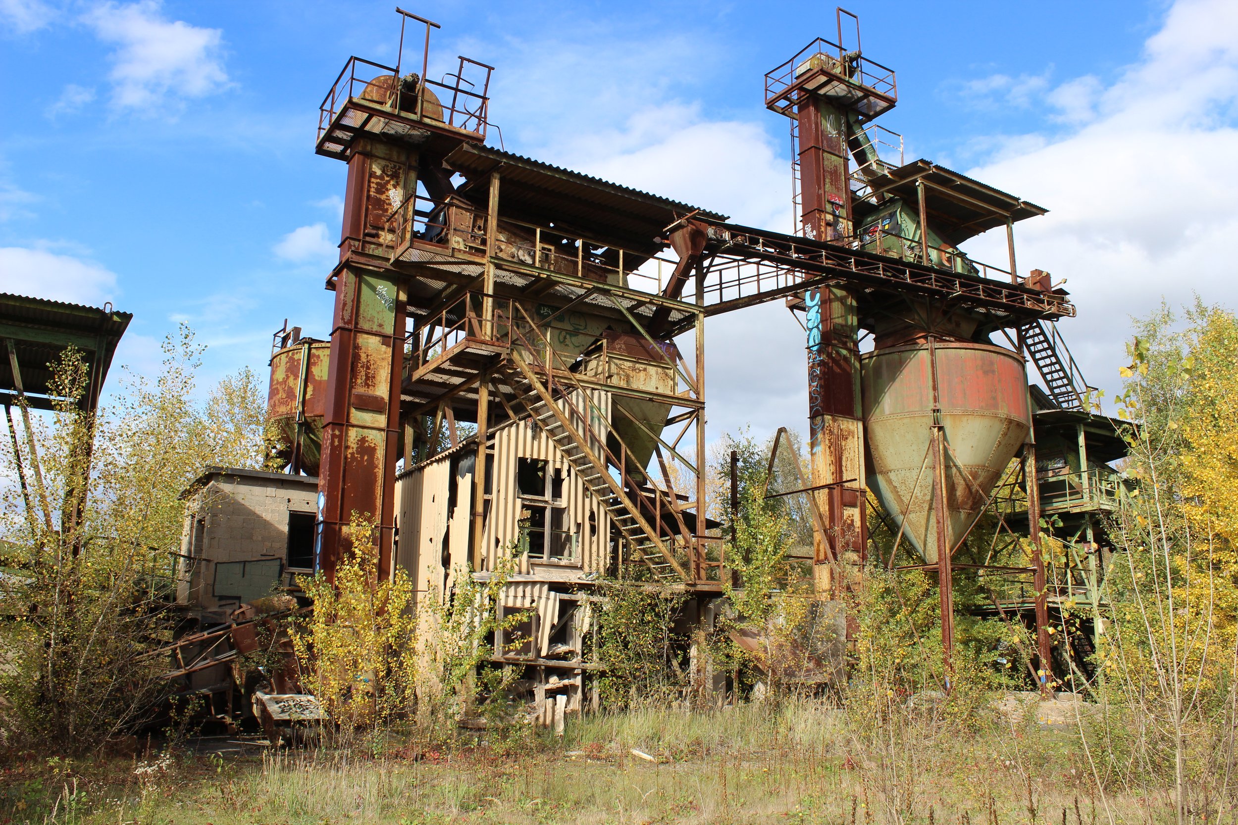 Verrostete, verlassene Industrieanlage mit Schornsteinen und Stufen, umgeben von Büschen und Bäumen, unter einem blauen Himmel mit Wolken.