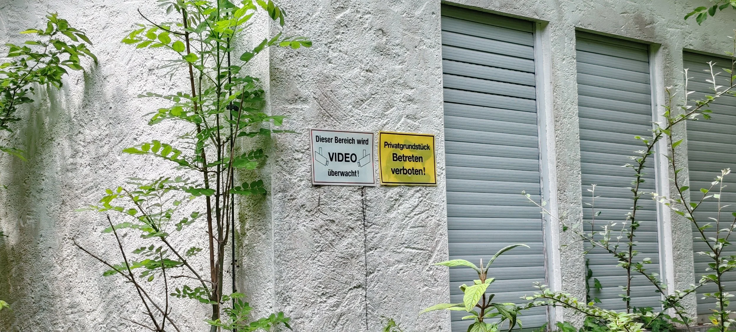 Zwei Schilder an einer weißen, texturierten Wand, die sagen: 'Dieser Bereich wird VIDEO überwacht!' und 'Privatgrundstück Betreten verboten!'. Umrandete offizielle Hinweise, um Privatsphäre und Überwachung zu betonen.