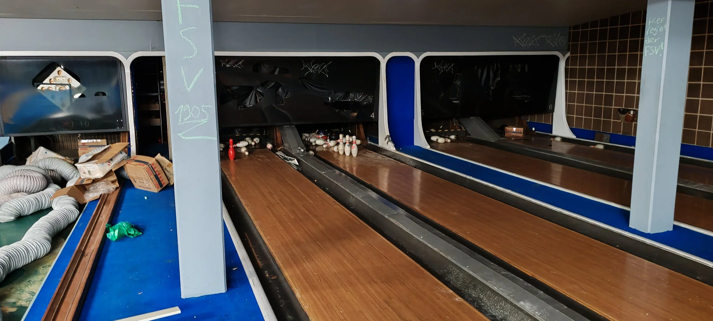 Leere Bowlingbahn mit 4 Bahnen, Bowlingkugeln, Pins und Zubehör. Graue und blaue Wände, Mitternachtsfarben, auf einer Seite von Verpackungsmaterial umgeben.