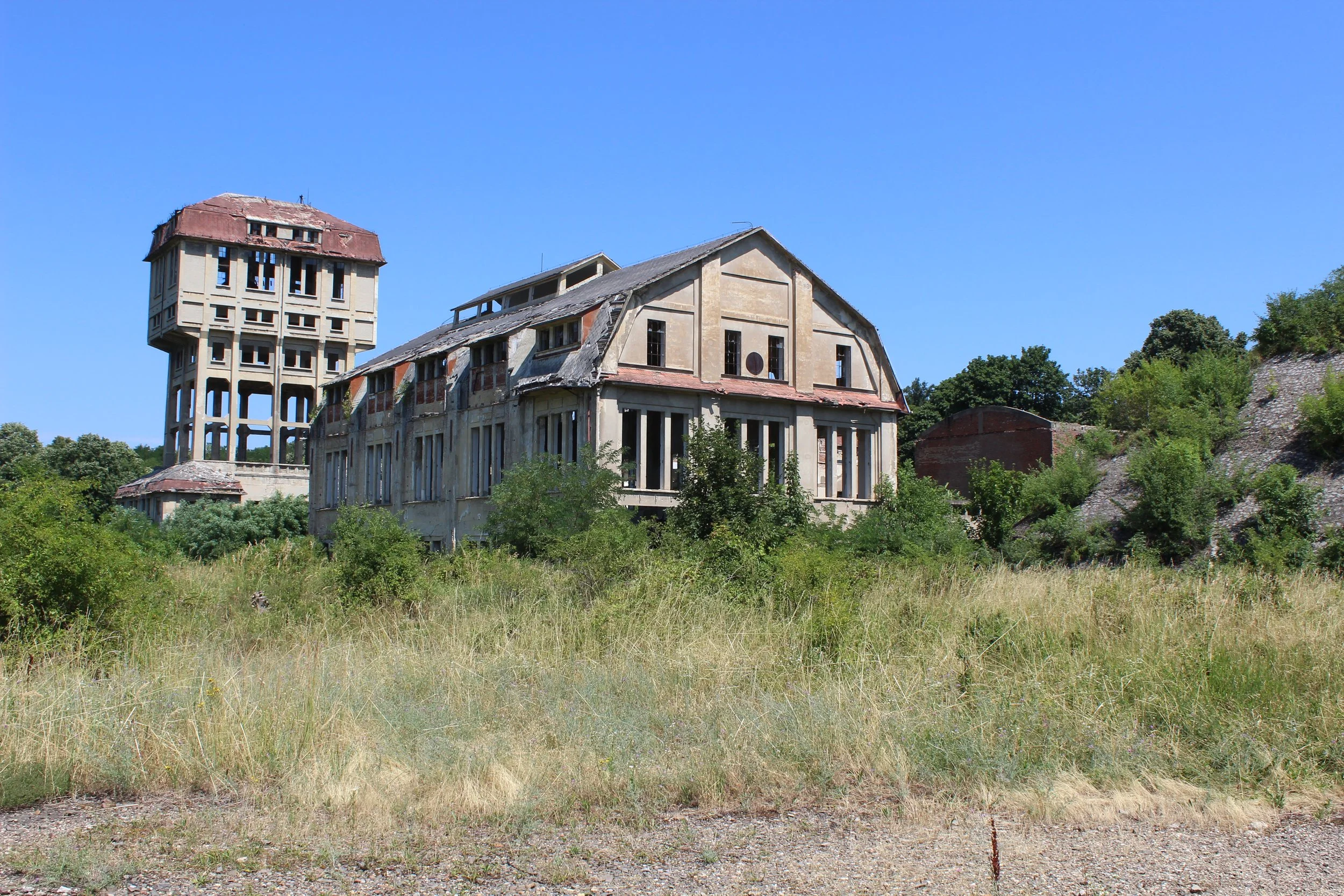 Verfallene Gebäude in einem grünen, bewaldeten Gebiet unter klarem blauen Himmel.