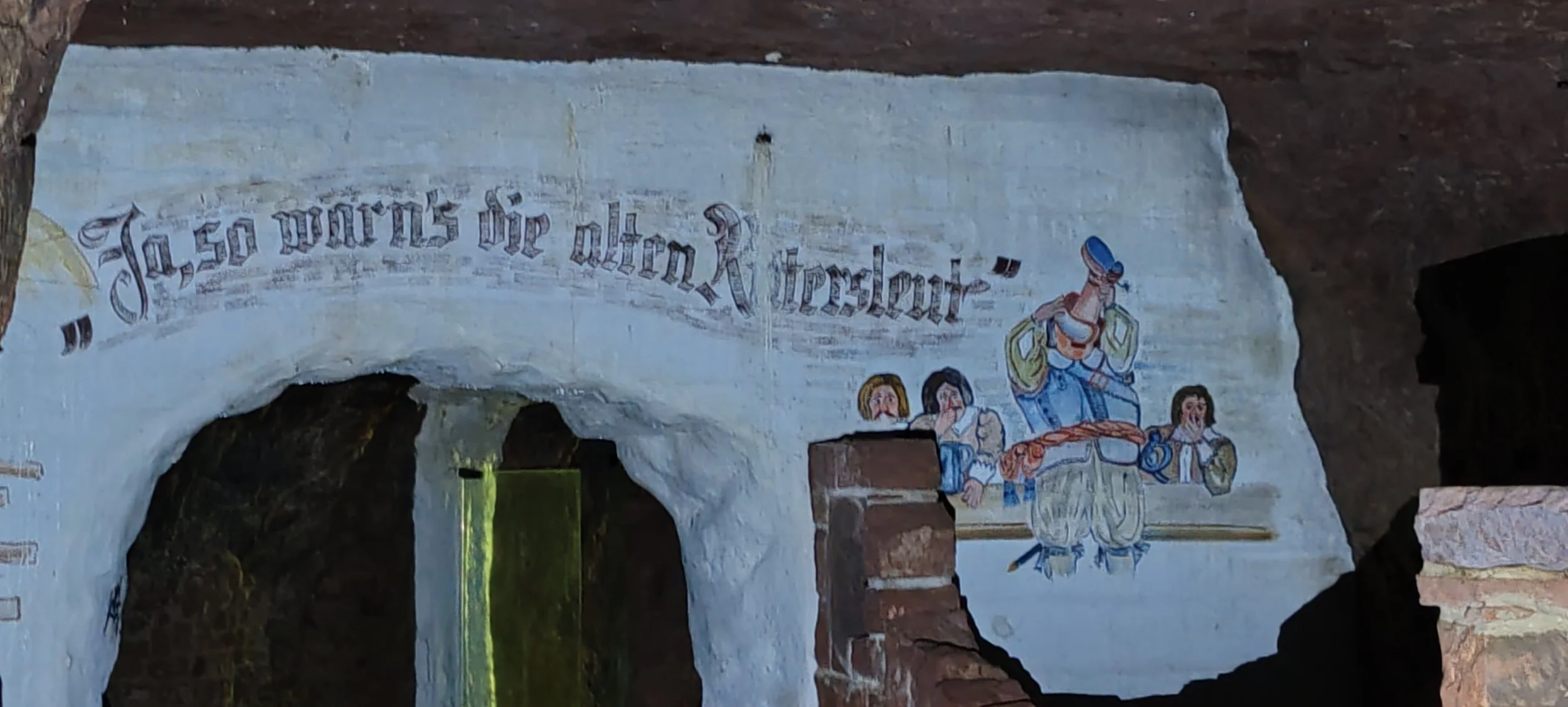 Handbemalte Wandbild in einer Höhle mit deutschen Schriftzug 'Haas wirft die alten Knochen' und einer Zeichnung von vier Menschen, drei Frauen und einem Mann in traditioneller Kleidung.