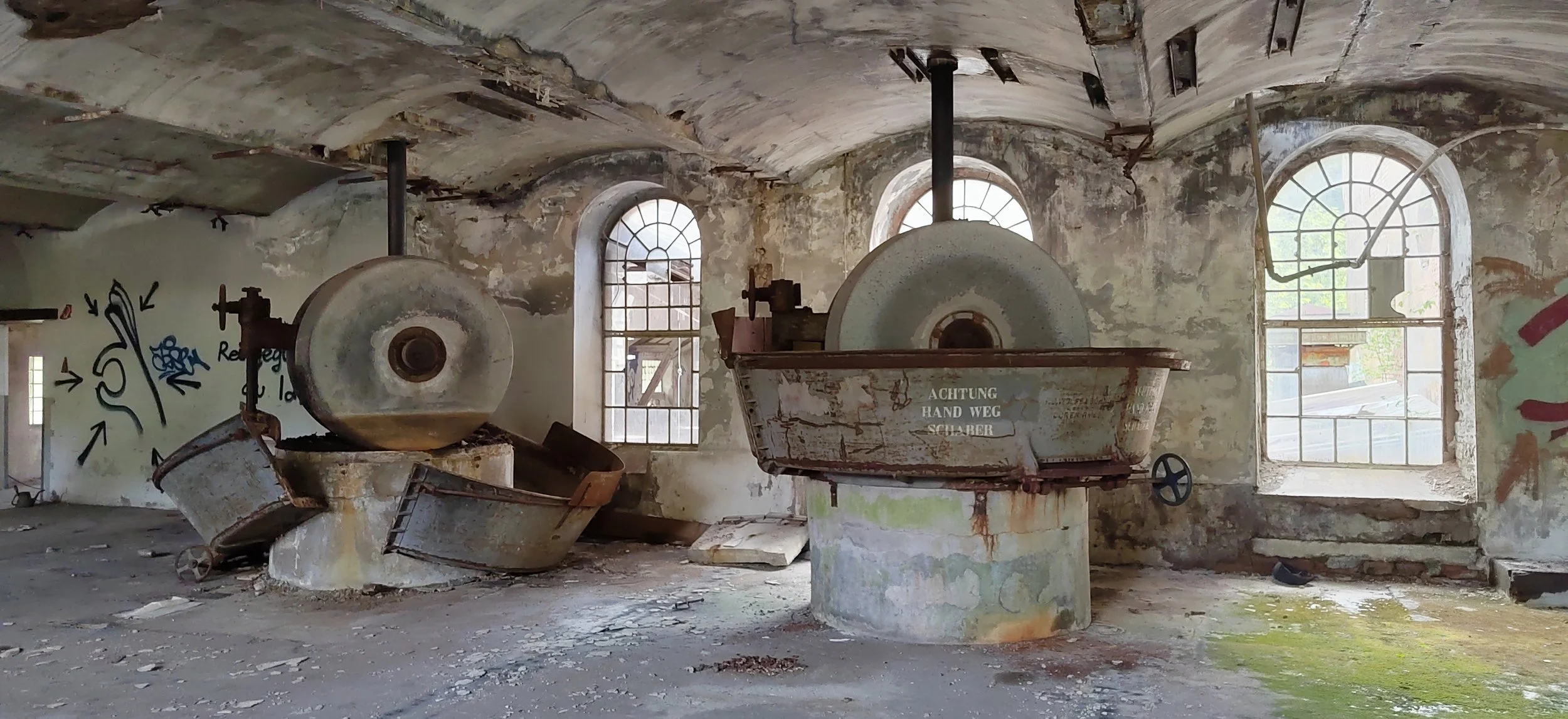 Verlassene industrielle Raum mit alten, rostigen Waschbecken, Wänden mit Graffiti, großen Fenstern mit Rost und Blick auf eine verfallene Umgebung im Hintergrund.