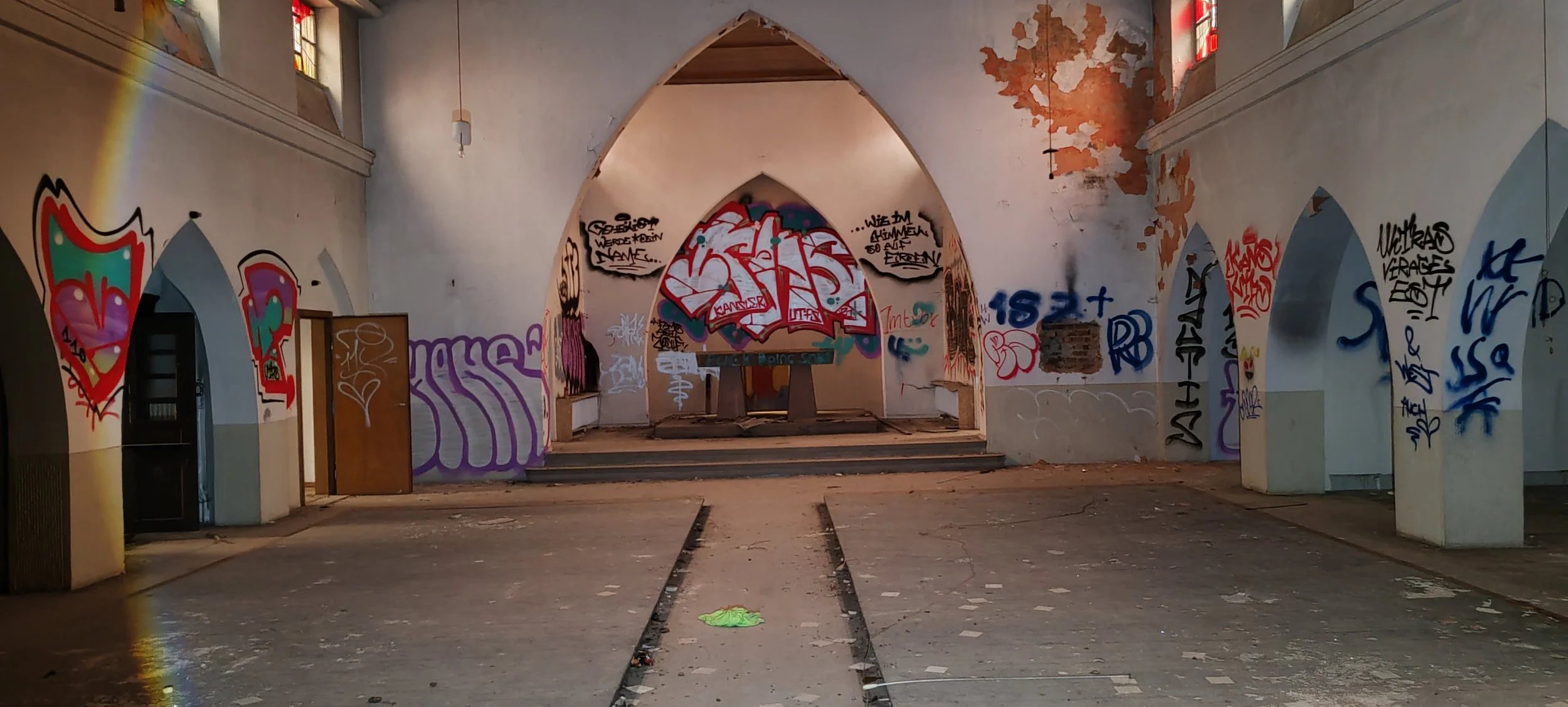 Innenraum eines verfallenen Gebäudes mit Graffiti an den Wänden, einem Altar im Hintergrund und bunten Fenstern.