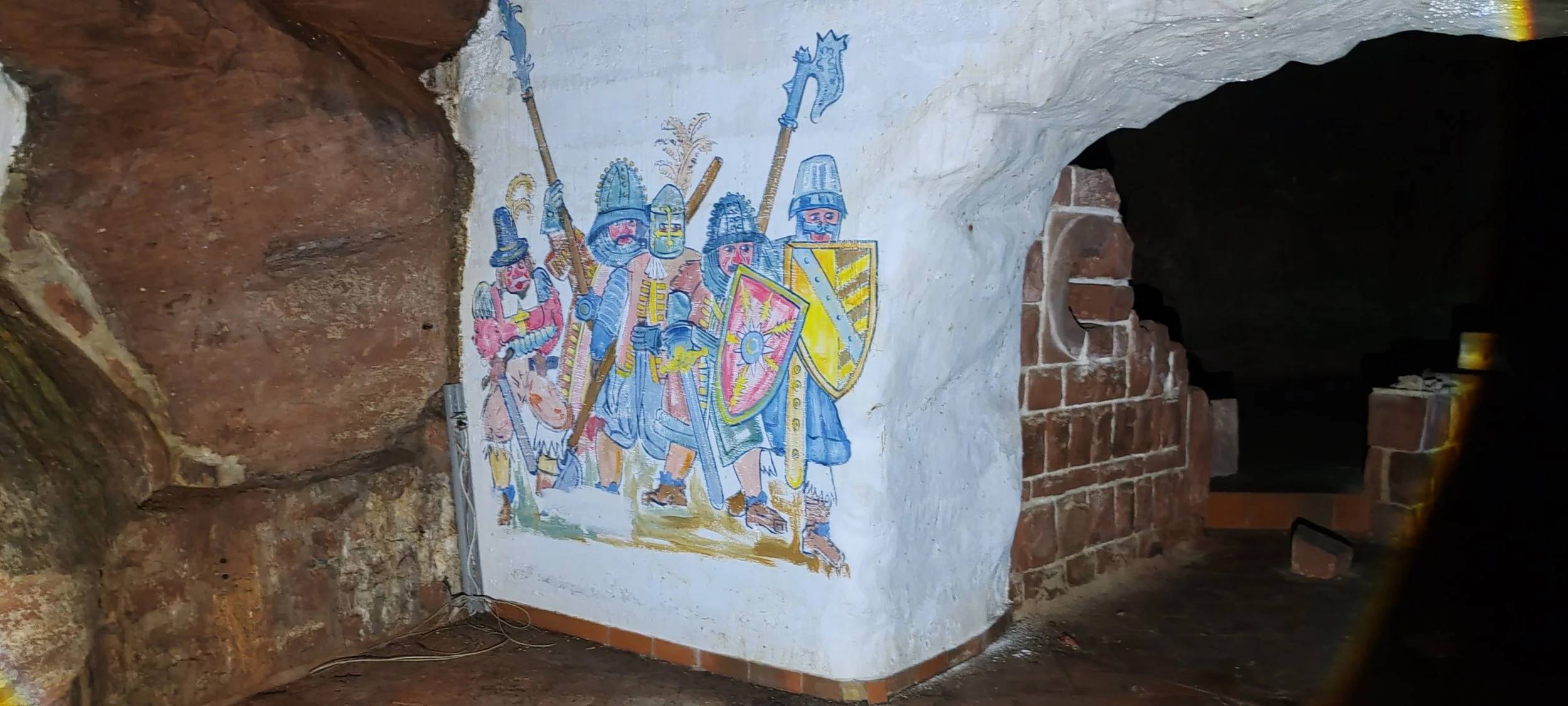 Wandmalerei mit mittelalterlichen Rittern mit Schwertern und Schilden, in einer dunklen, brick- und steinverzierten Umgebung.