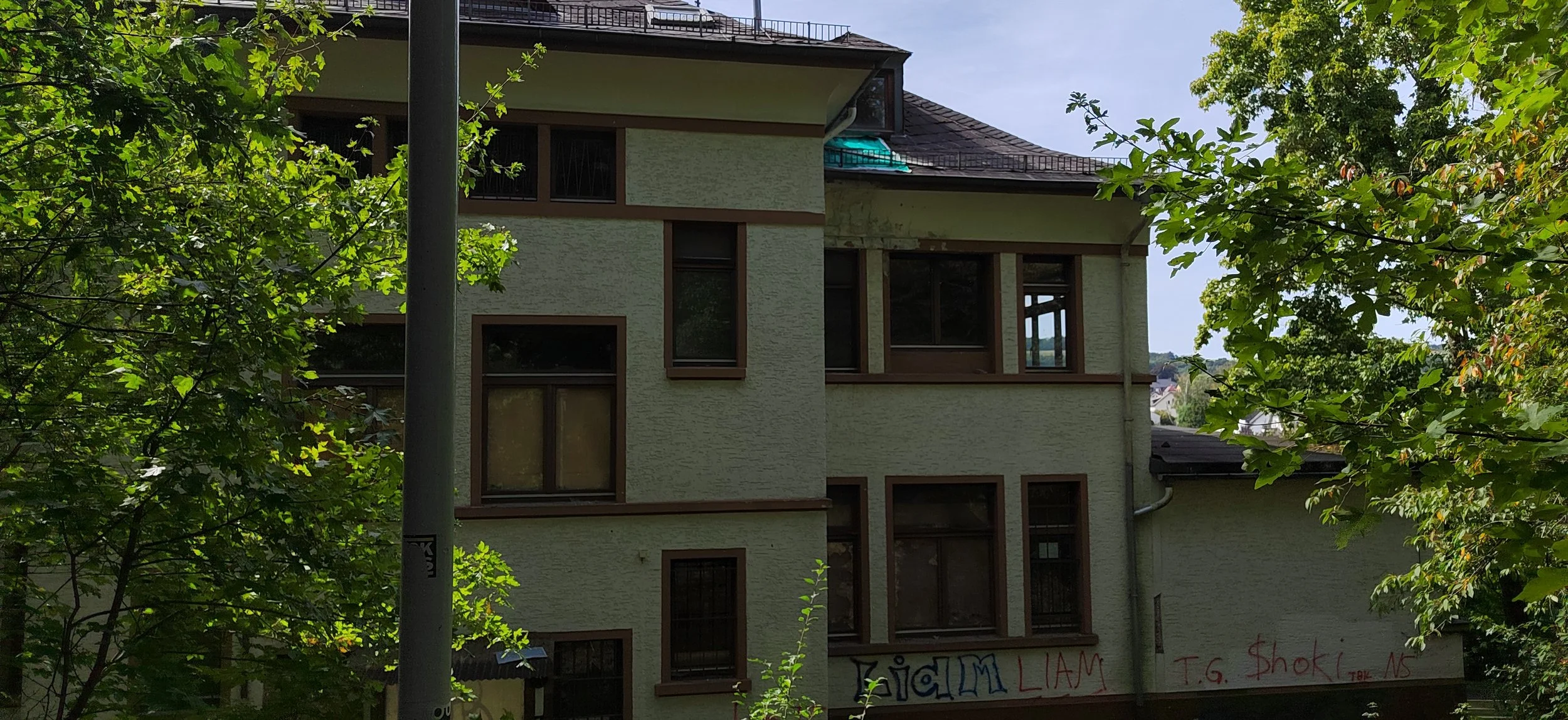 Mehrstöckiges Gebäude mit mehreren Fenstern, umgeben von grünen Bäumen, mit Graffiti an der Wand. Im Vordergrund ein Baum und eine Straßenlaterne.