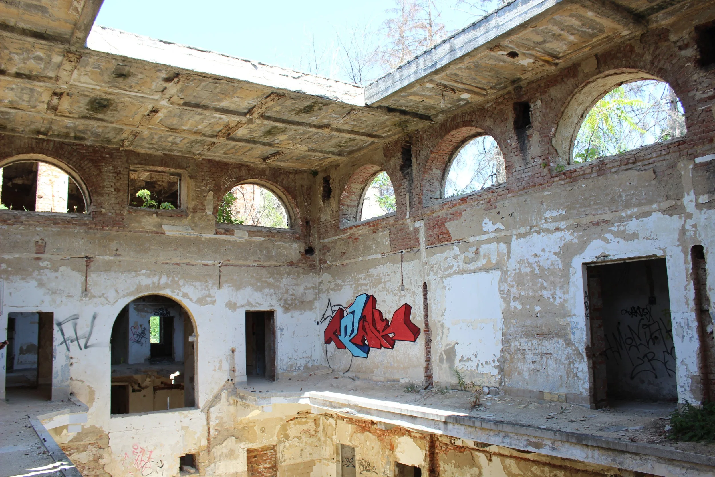 Ein verfallenes Gebäude mit durchbrochenen Wänden und mehreren Bögen. Graffiti auf der Wand im Vordergrund. Das Dach ist teils zerstört, und es sind Bäume im Hintergrund sichtbar.