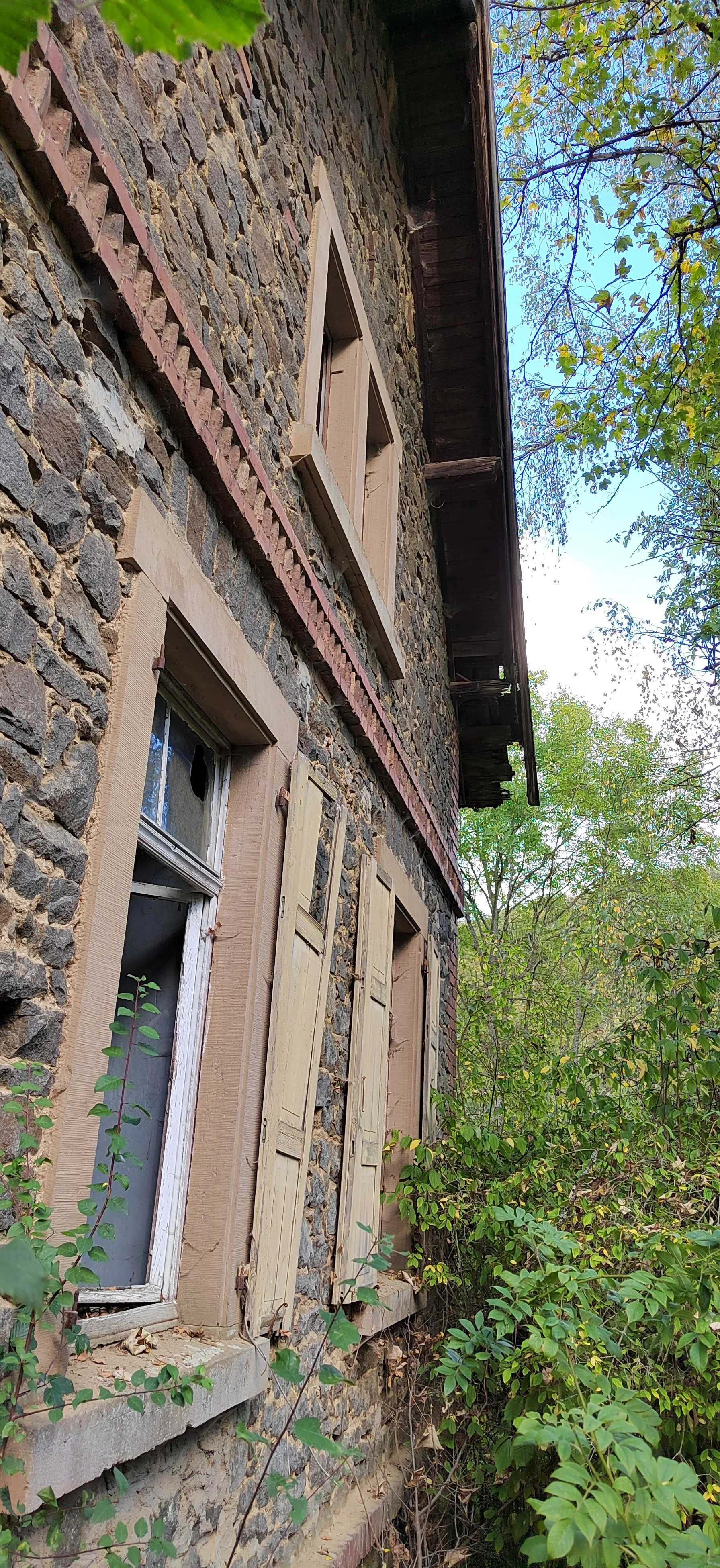 Ein altes Steinhaus mit Fenstern und Holzzierläden, umgeben von Büschen und Bäumen, mit einem blauen Himmel im Hintergrund.