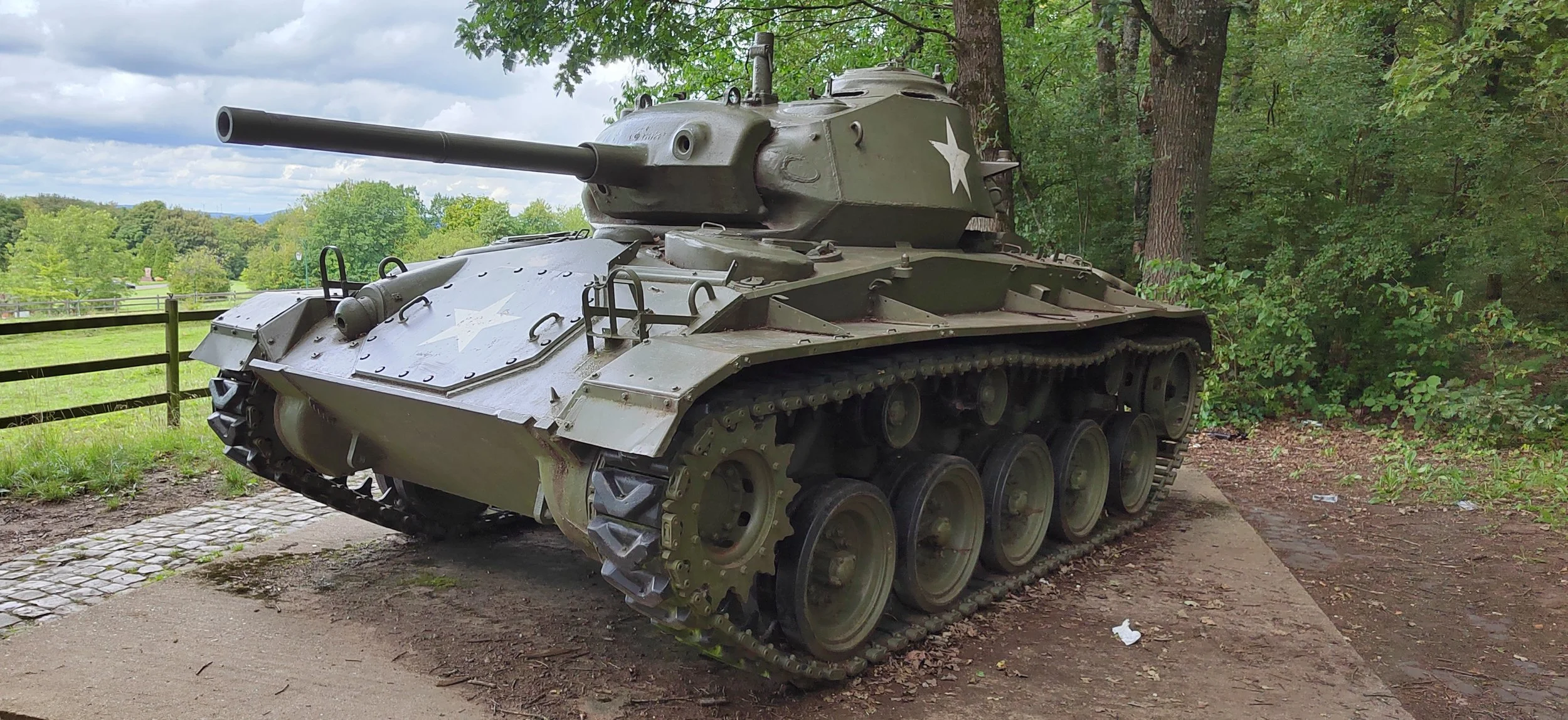 Ein alter Grauer Panzermaschine mit Sternschild auf dem Turm, steht auf einem Bett aus Erde und Steinen im Grünen, umgeben von Bäumen und Himmelswolken.