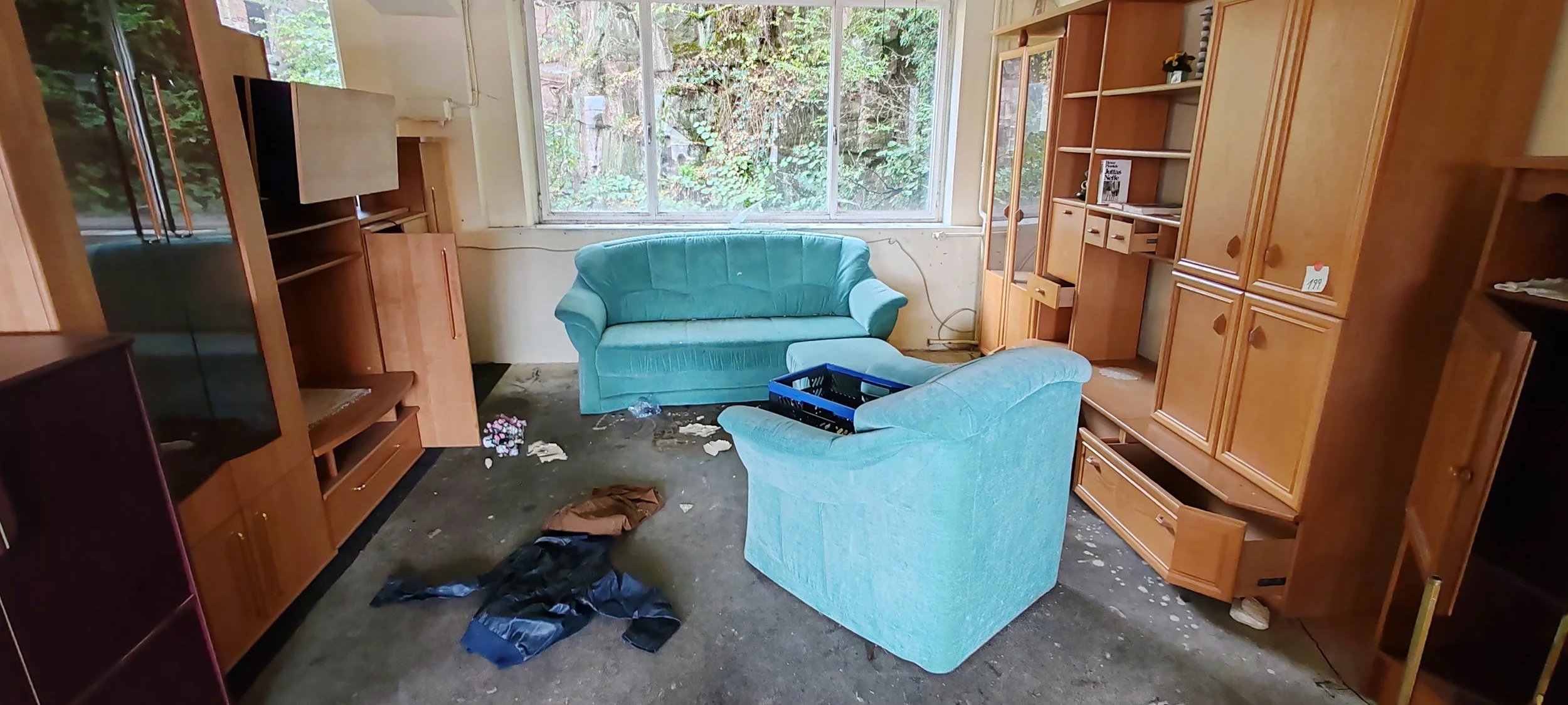 Verlassener Wohnraum mit zwei blauen Sofas, unordentlichem Boden und großen Fenster mit Blick auf einen Garten im Hintergrund.